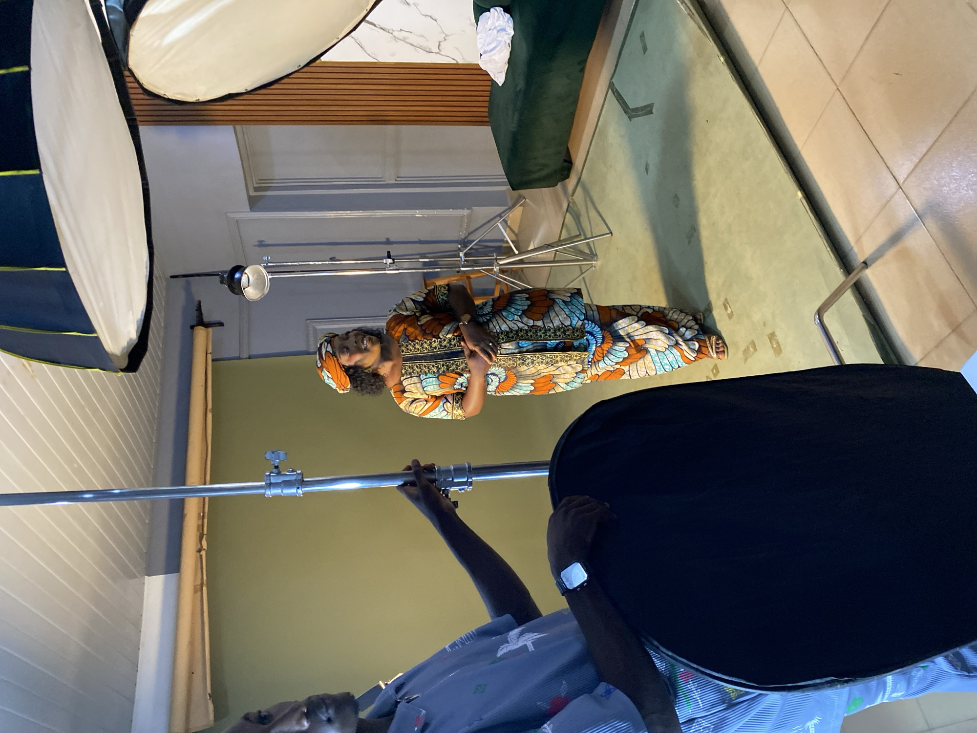 This image, rotated 90 degrees clockwise, captures an indoor photography or video shoot in Bukuru, Nigeria.
**Subjects:** The central figure is a woman, likely a model, dressed in a striking, multi-colored African print (Ankara) dress with gold accents and a matching headwrap. She stands with a gentle smile, her arms crossed in front of her. In the foreground, a crew member is partially visible from the chest down, holding a large, black circular reflector or diffuser. They are wearing a light blue striped shirt and a watch on their left wrist.
**Scene and Setting:** The event unfolds within an indoor space, possibly a studio or a styled room. The background features a light olive-green wall, above which is a section of wooden slats and a white, marble-patterned panel. A dark green sofa with a white crumpled cloth is positioned in the upper right. The floor is tiled in a mix of light green/grey and beige sections, and the ceiling is white and paneled.
**Objects:** Professional photographic equipment dominates the setup. Two large, white softboxes or octaboxes are prominent on the left, serving as key light sources. Multiple metal light stands are present, with one supporting a smaller, spotlight-like fixture behind the woman. The crew member actively positions a black reflector/diffuser, suggesting light shaping.
**Activity:** The image depicts a moment during a professional portrait session or media production, with the woman posing under controlled lighting conditions, managed by the visible crew member and equipment.
**Notable Details:** The woman's traditional attire is a vibrant highlight. The artificial lighting setup indicates a deliberate and controlled environment for the shoot, making the actual time of day less relevant to the scene. There is no visible text within the frame.