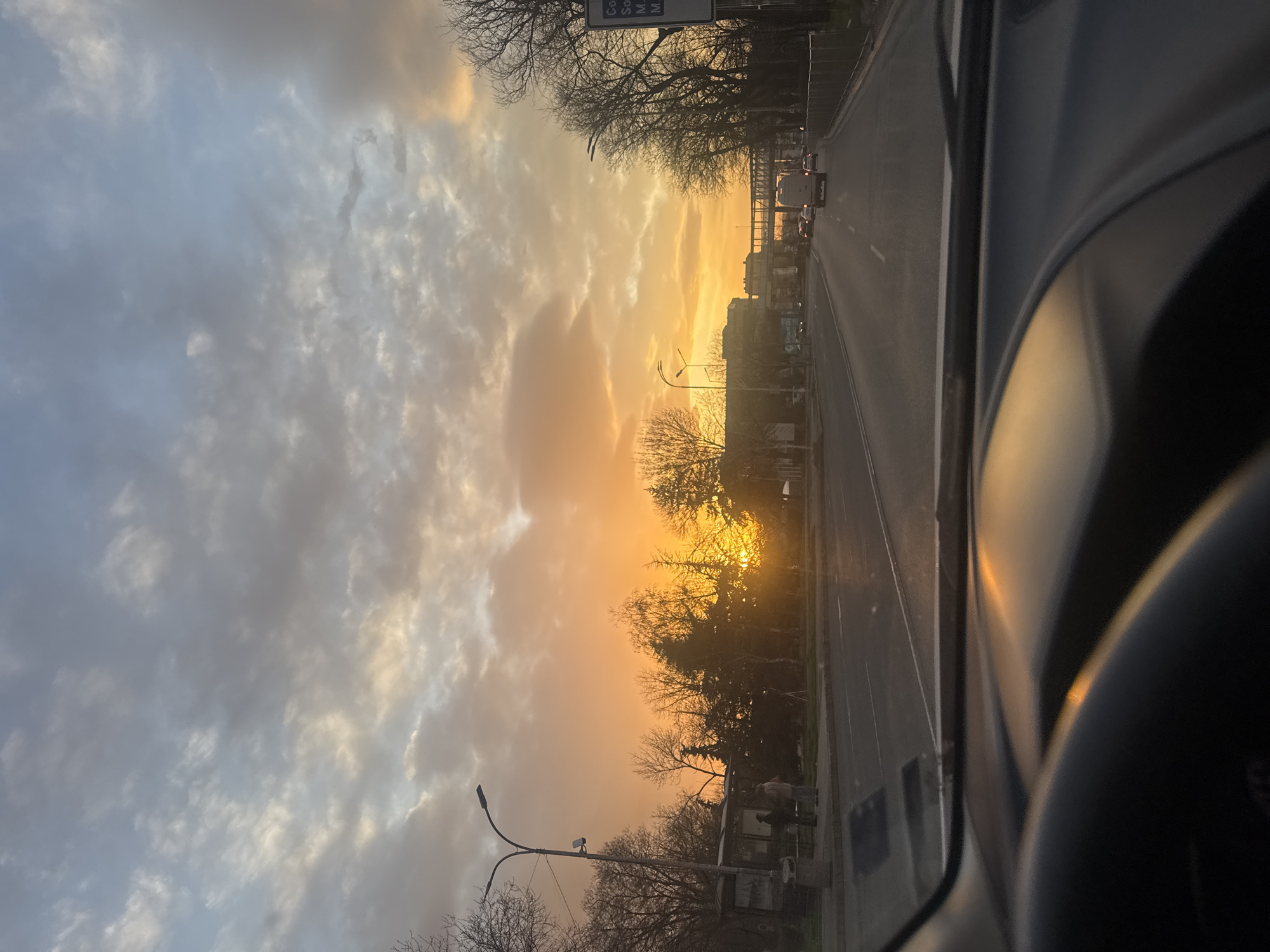 The photograph, taken from inside a vehicle moving along a road, captures a sunset scene. In the middle ground, the sun appears to be setting directly ahead down the road, its light partially obscured by trees and buildings lining the route. A railway bridge is also visible. The sky is a mix of grey clouds and orange light from the setting sun. A few vehicles can be seen on the road ahead. Elements of the car's interior, including the dashboard and steering wheel, are visible in the foreground. A bus station is on the side of the road. Based on visual clues such as the architecture and plant life, the location may be Burgas, Bulgaria.