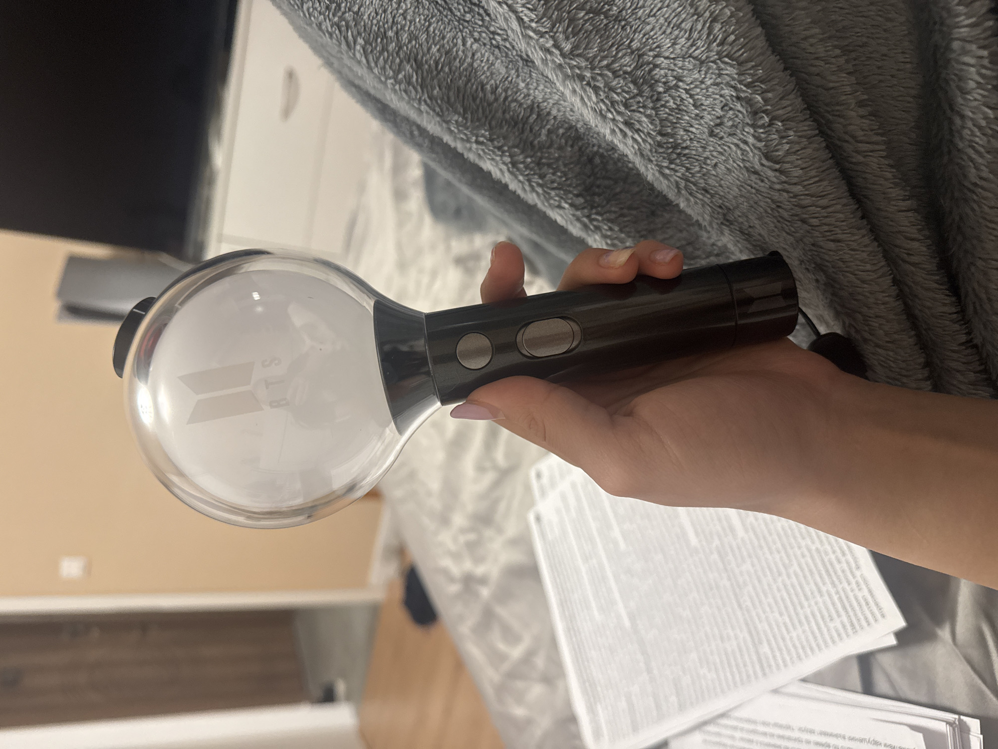 The image depicts a close-up, first-person perspective of a right hand holding a BTS "ARMY Bomb" light stick indoors. The light stick features a clear spherical dome with the illuminated white BTS logo and text inside, attached to a dark, matte handle with two circular, light grey buttons. A dark lanyard or strap extends from the base of the handle. The person's fingernails are visible, with light pink polish on at least one finger.
To the left of the hand, a stack of white papers with dense printed text, appearing to be in a Cyrillic script, rests on a light-colored surface with a subtle geometric pattern, possibly a bedspread or blanket. A grey, fluffy, textured blanket or throw is visible in the lower right and midground, partially covering the patterned surface.
In the background on the left, a plain, light beige wall is visible, with a white rectangular object (likely a light switch or outlet). A partially open wooden doorway leads into another room, where a dark object is on the floor. In the background on the right, a large, dark rectangular screen, presumably a television, is positioned above a piece of white furniture. To the left of the screen, a tall, light-colored vertical object, consistent with a gaming console (e.g., PlayStation 5), stands on the white furniture. The scene is illuminated by indoor ambient lighting.
