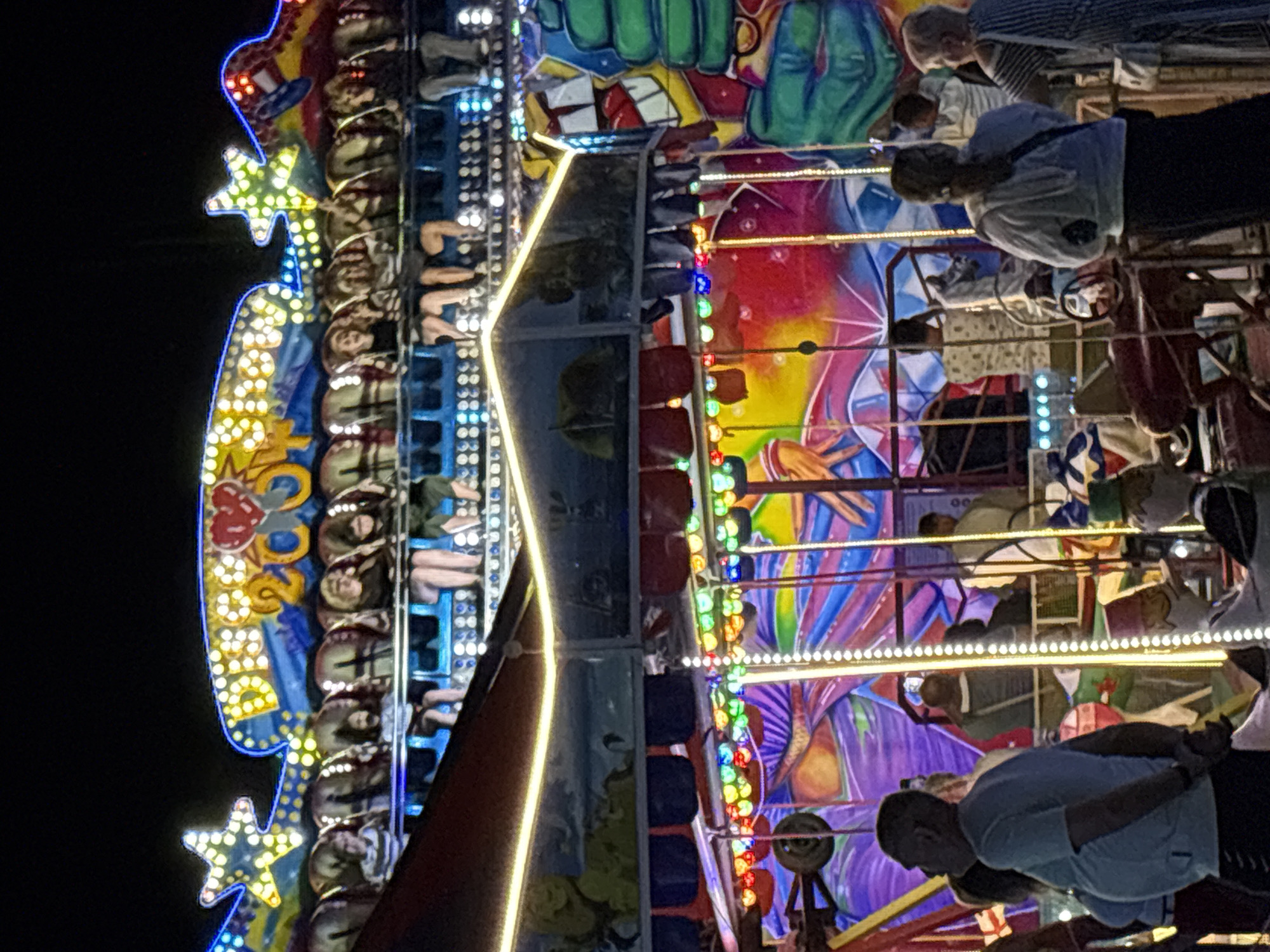 The image depicts a brightly lit amusement park ride at night. The ride features multiple levels with various seating arrangements for participants, some of whom are visible. The ride is adorned with numerous colorful lights, including flashing bulbs and illuminated signs, one of which appears to read "DISCO 2000" with a heart symbol. The background is a dark night sky, contrasting with the vibrant colors of the ride and its decorations. The ride itself appears to have a carnival-like aesthetic, with painted murals and whimsical figures visible. The overall scene suggests a lively atmosphere at an amusement park.