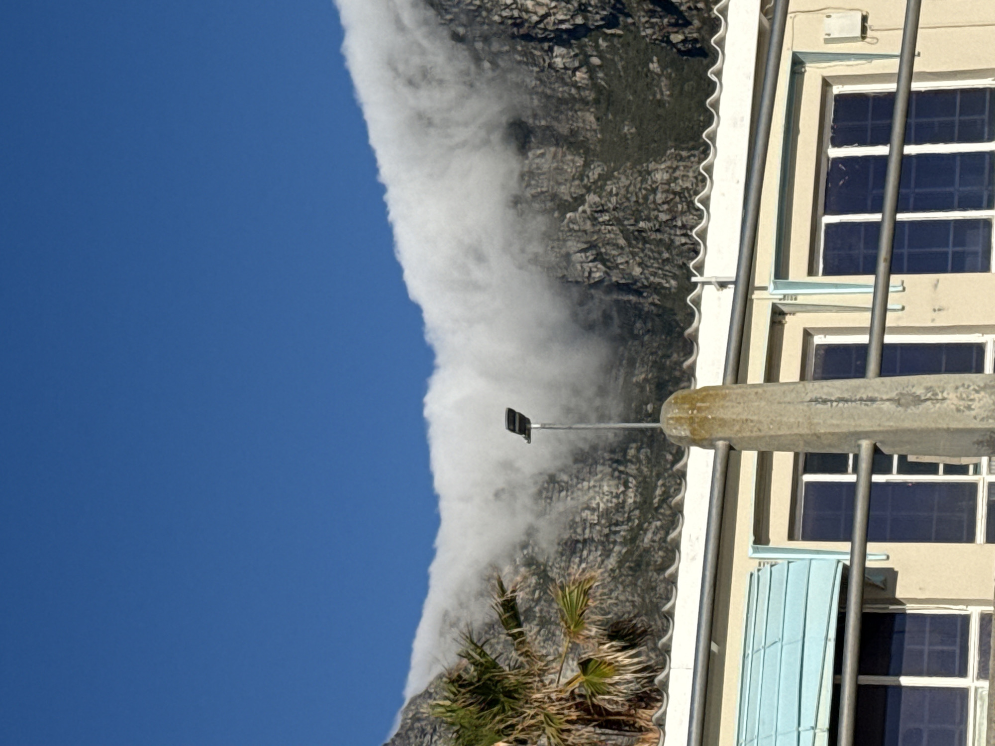 The image depicts a landscape featuring Table Mountain in Cape Town, South Africa, shrouded in a layer of cloud cover extending down its slopes under a clear blue sky. A building with several windows is visible on the right side of the frame, along with a street lamp. Some green plants are seen near the base of the mountain.