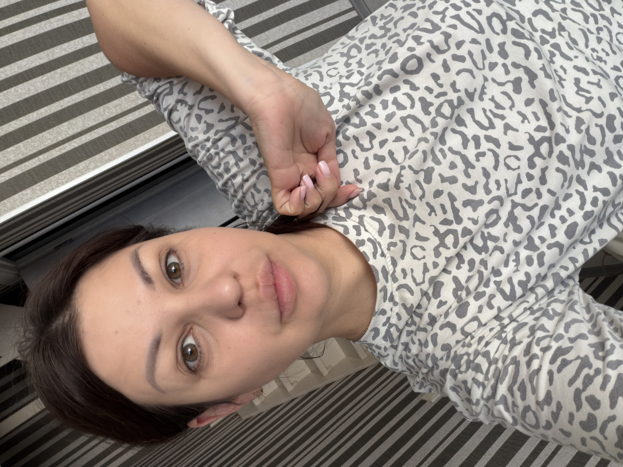 This is a medium close-up, selfie-style photograph of a woman lying down. She is wearing a grey and white leopard print top. Her left arm is bent at the elbow and her hand rests on her chest, with her fingernails painted a light pink. The background consists of a striped surface, appearing to be a bed or sofa, with alternating dark grey and light grey stripes.
The woman has dark brown hair and her head is tilted. She has brown eyes and is looking towards the camera. Her skin appears smooth and she is wearing light makeup. Her lips are slightly parted. The overall lighting suggests it is daytime, possibly indoors near a window. The image has a casual and relaxed feel, implying the woman is at home. No text is visible in the image. The context provided indicates the location is Sochi, Russia.
