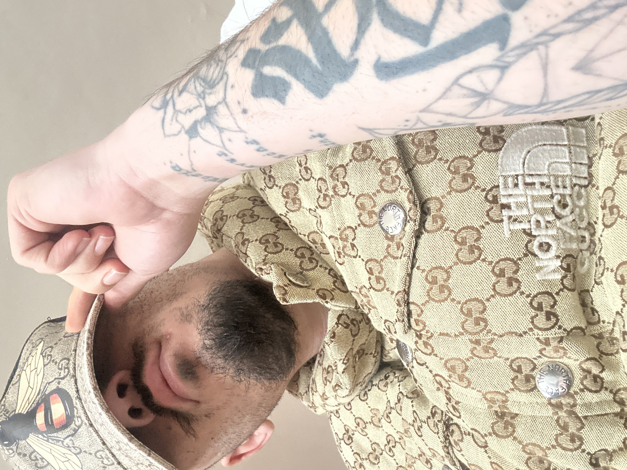 The image displays a close-up, low-angle shot of a male individual, seen from the mid-chest upwards, situated in Varna, Bulgaria. The individual wears a tan baseball cap adorned with a repeating 'GG' monogram pattern and a prominent multi-colored bee emblem on the front. His left hand is raised, with fingers curled, resting against the cap's brim, partially obscuring his forehead. Visible facial features include a mustache and a short beard. He is dressed in a tan garment, consistent with a jacket, also featuring the 'GG' monogram pattern. This garment is equipped with silver-colored circular buttons, some embossed with "THE NORTH FACE," and an embroidered light-colored "THE NORTH FACE" logo prominently displayed on the chest area. His visible left forearm is extensively covered with tattoos, including dark stylized characters and intricate linear designs. The background is an undifferentiated, light-colored surface.