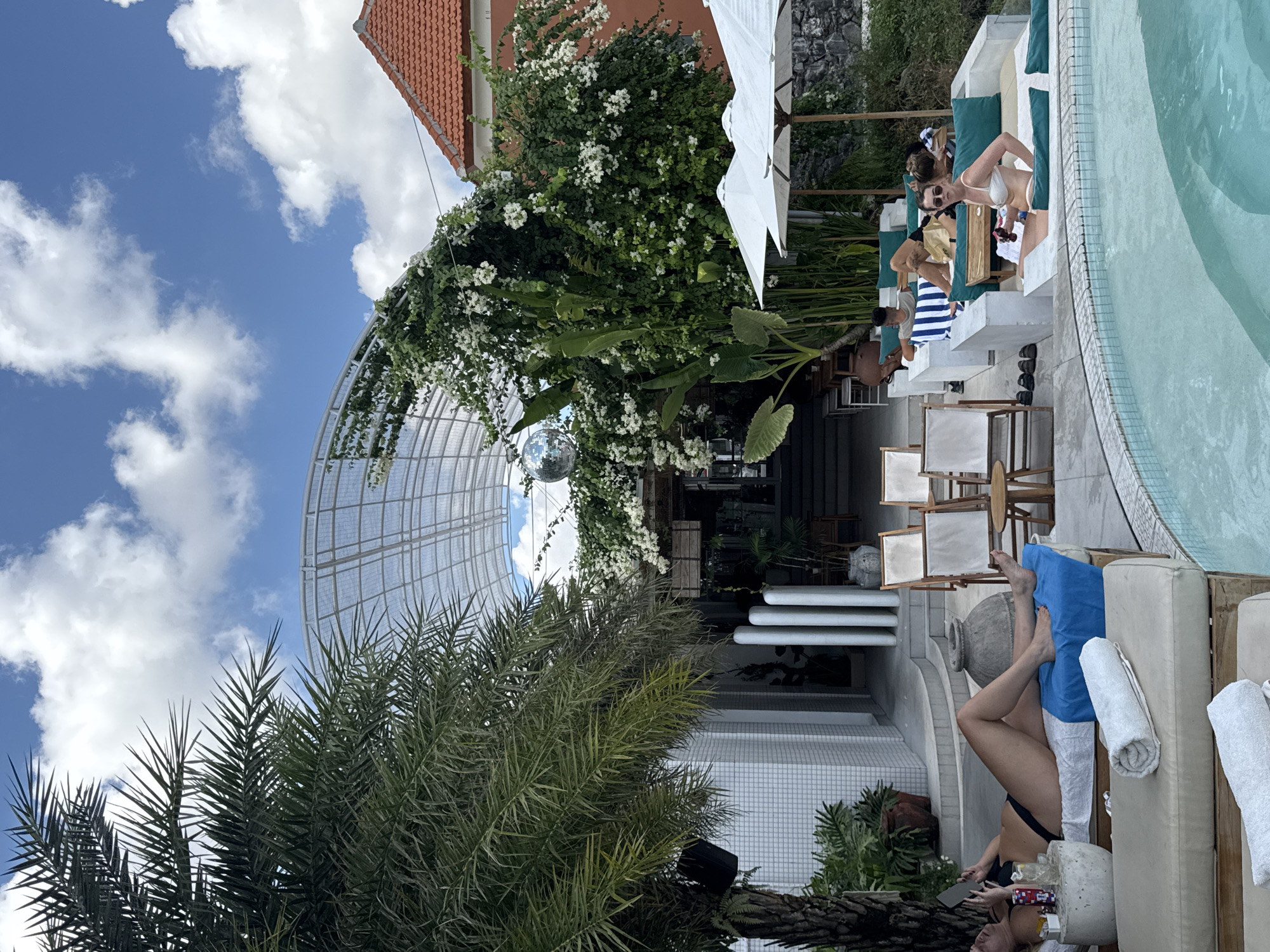 This image depicts a vibrant outdoor leisure area, likely a resort or beach club in North Kuta, Indonesia, bathed in bright daylight under a blue sky with scattered white clouds. The scene is dominated by a swimming pool and various relaxation spots, surrounded by lush tropical greenery and modern architectural elements.
In the foreground, a person in a black bikini is relaxing on a light-colored poolside lounger, holding a smartphone. A blue towel and white rolled towels are visible next to them. Further along the pool, a woman in a white bikini and sunglasses sits upright on a lounger, looking to the side. Other people are seen relaxing on loungers around the clear, light blue pool, some partially obscured by white patio umbrellas.
The left side of the image features abundant tropical plants, including large palm fronds. A distinctive arched, grid-like structure, possibly a conservatory or walkway roof, is heavily adorned with white flowering vines, from which a reflective disco ball hangs. Behind the vegetation, a modern building with white tiled walls and cylindrical columns is visible, along with white stairs. In the background, a building with a reddish-brown tiled roof and a stone wall are partially visible amidst more greenery.
The time of day appears to be midday or early afternoon due to the bright, direct sunlight. The overall setting suggests a luxurious and relaxing tropical vacation spot. There is no clearly legible text visible in the media file.