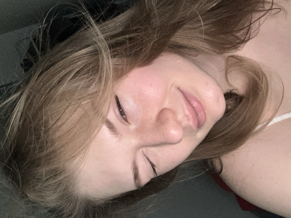 A woman with light brown hair and fair skin is lying down, her head turned to the right. Her eyes are partially closed, and she is smiling slightly. Her lips are full and pink. The background is dark and out of focus, with a hint of gray and black, possibly from clothing or bedding. A thin white strap is visible on her shoulder, suggesting a tank top or camisole. The lighting is soft and muted.