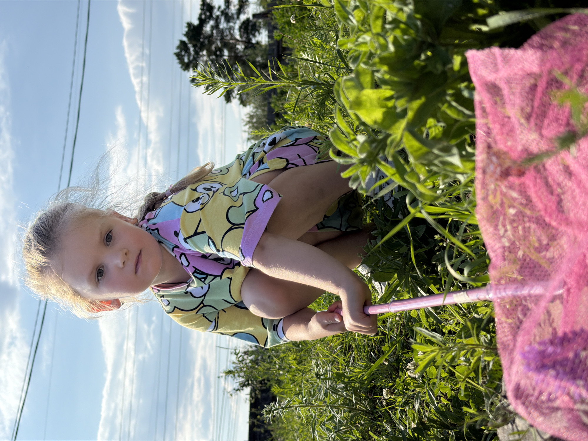 A young girl is crouched in tall green grass, holding a pink butterfly net with a purple net. She is wearing a yellow t-shirt with a colorful cartoon print of characters with large round ears and eyes. Her blonde hair is pulled back, and she is looking directly at the camera with a neutral expression. The sun is shining, casting shadows on the grass and illuminating the girl's face and hair. The sky in the background is blue with scattered white clouds. In the distance, there are trees and what appear to be power lines. The scene suggests a sunny day outdoors, possibly in a rural or park setting. The image is oriented vertically, as if taken from a low angle.