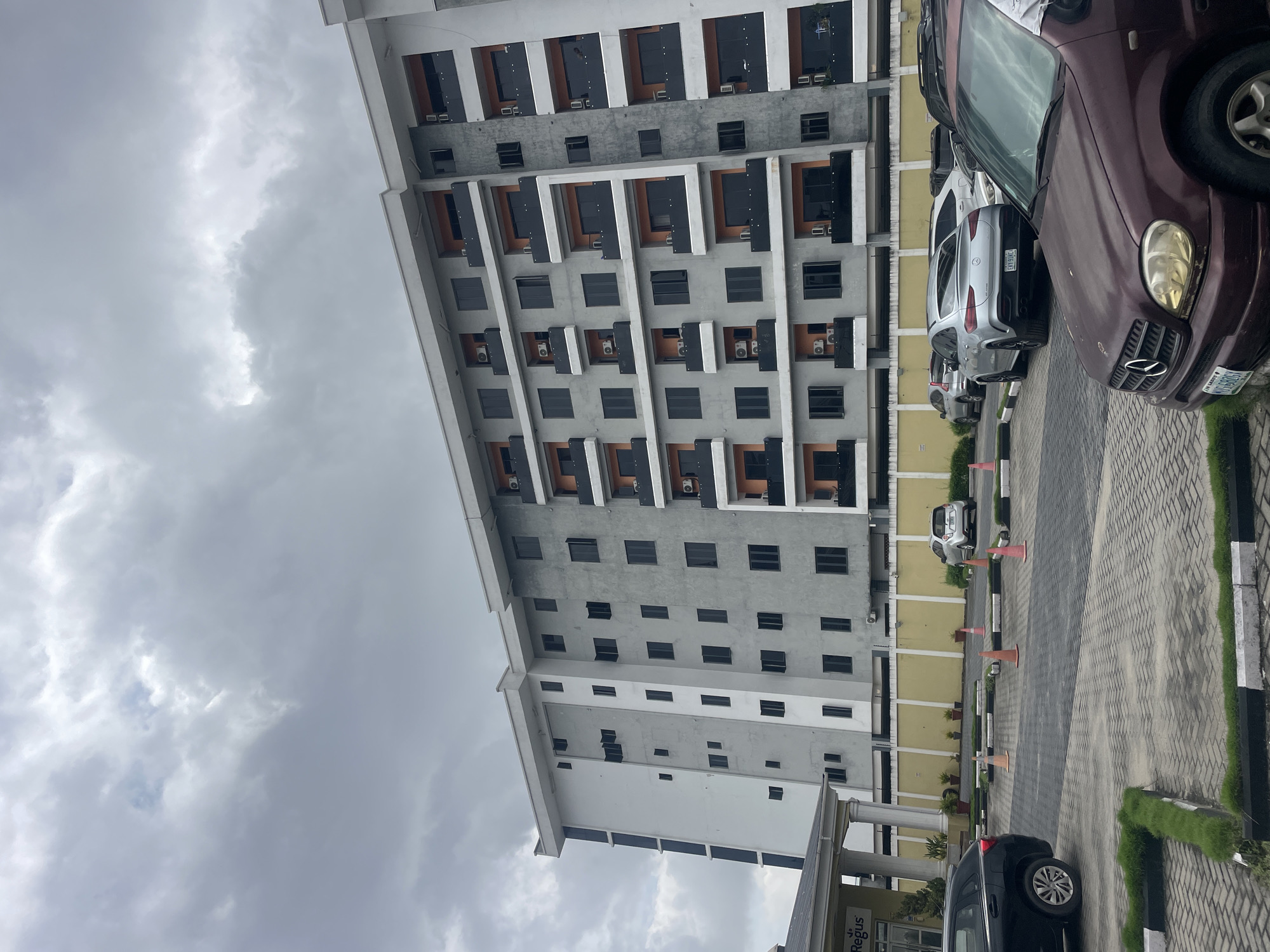 In Moba, Nigeria, a modern apartment building with multiple stories stands under a cloudy sky. The building features a grey facade with white accents and numerous dark-tinted windows and balconies. Several air conditioning units are visible on the exterior walls. In the foreground, a paved parking lot is marked with white lines and dotted with orange traffic cones. A dark-colored car is parked on the left, and a silver coupe with the license plate "CRY 918L" is parked towards the center. A dark red SUV is in the immediate foreground, partially obscuring the view of the parking lot. A low yellow wall runs along the back of the parking area, behind which several potted plants are arranged. The overall scene depicts an urban environment on an overcast day, focusing on the architecture and the parking area.