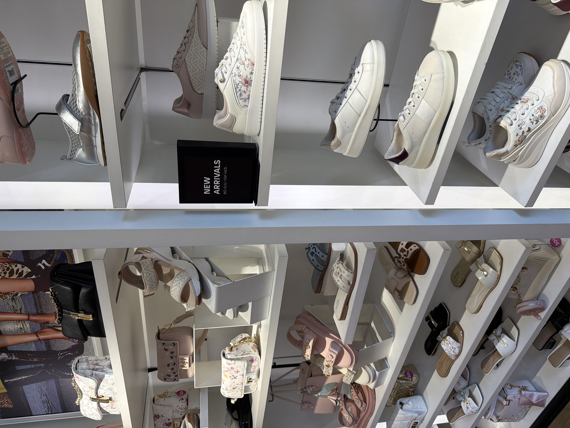 The image shows a retail display, likely in a store selling shoes and handbags. The shelves are white, displaying a variety of footwear, including sneakers and sandals, and handbags in various styles and colors. A black sign with "NEW ARRIVALS" is visible. The location is Nha Trang Ward, Vietnam.