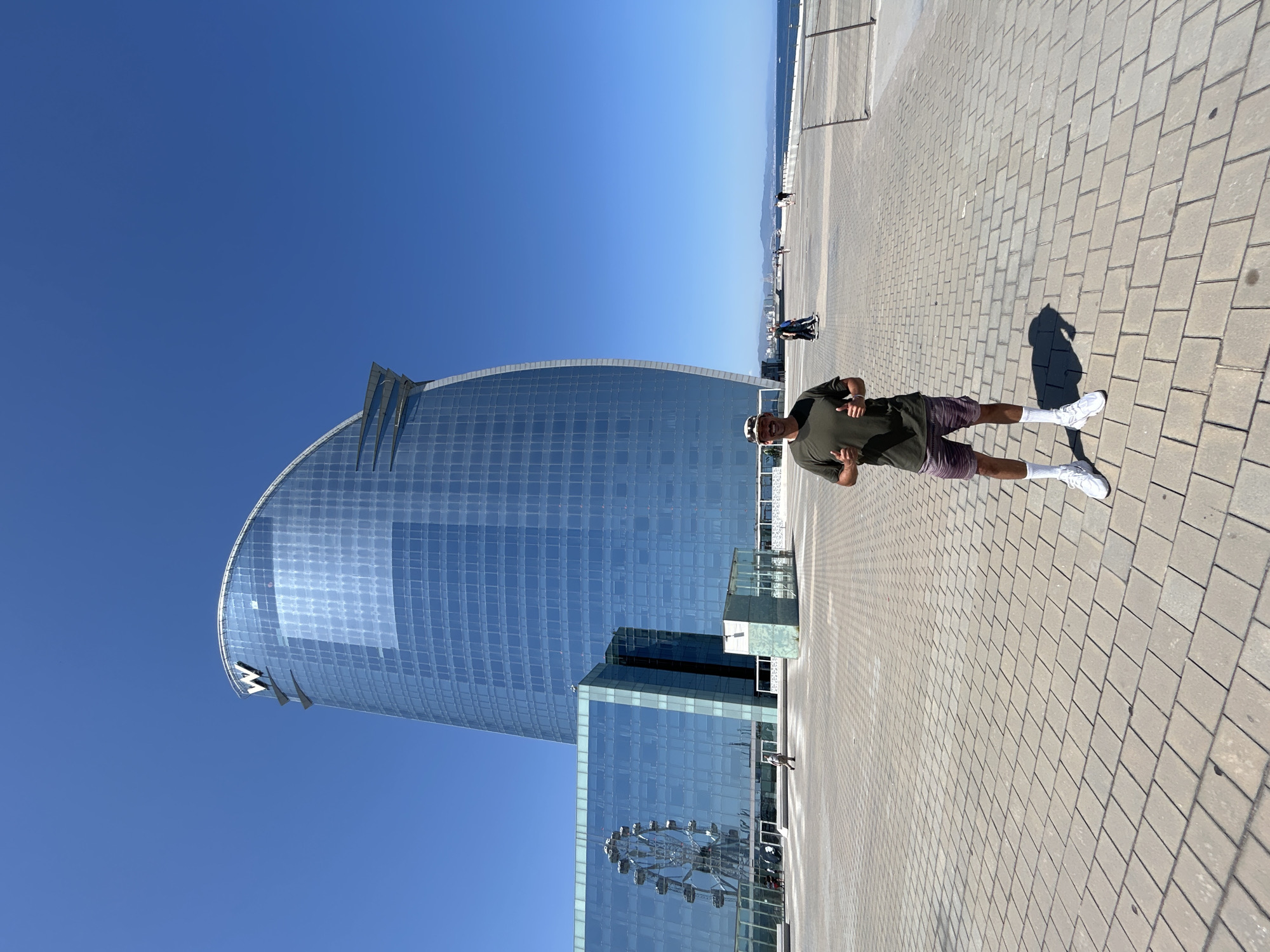 In this full-body, outdoor shot, a man stands in front of the iconic W Barcelona hotel, also known as Hotel Vela, on a clear, sunny day. The man, wearing a green t-shirt, patterned shorts, white socks, and white sneakers, is giving a thumbs-up with both hands. He is positioned on a wide, paved plaza, which features diamond-patterned stone tiling.
Behind the man, the W Barcelona hotel dominates the background with its distinctive sail-like glass facade. To the left of the hotel, a section of the building reflects a Ferris wheel. Other people can be seen walking in the distance on the plaza and along the waterfront. The sea is visible on the right side of the image, with a distant coastline and mountains on the horizon. The sky is a vibrant, cloudless blue.