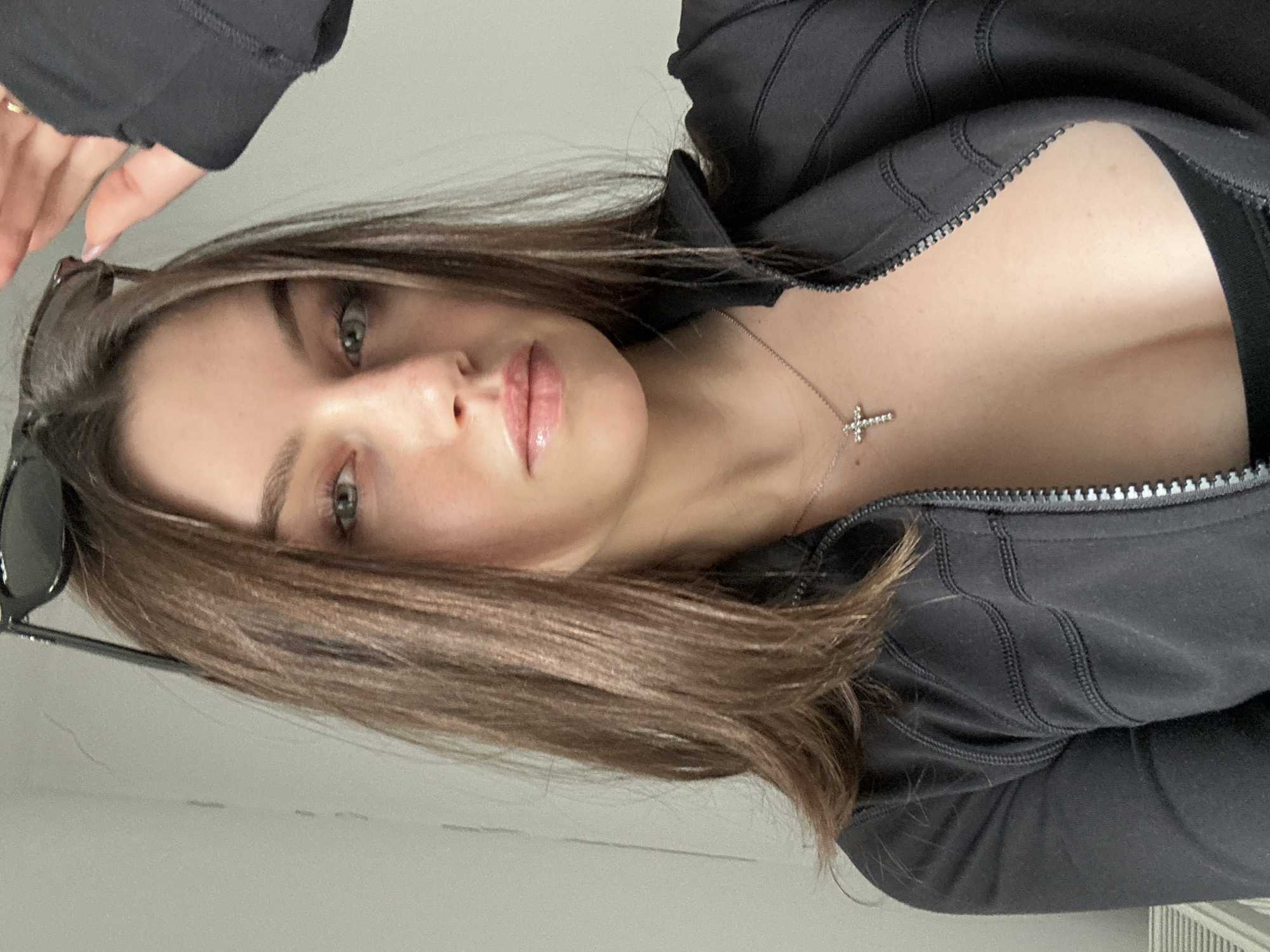 A selfie of a woman with brown hair and green eyes is angled to the right. She wears a black zippered jacket, and a silver cross necklace is visible. A pair of sunglasses is in the top left corner of the frame. The background appears to be a plain, light-colored wall. The location in the city is Unknown, Unknown.