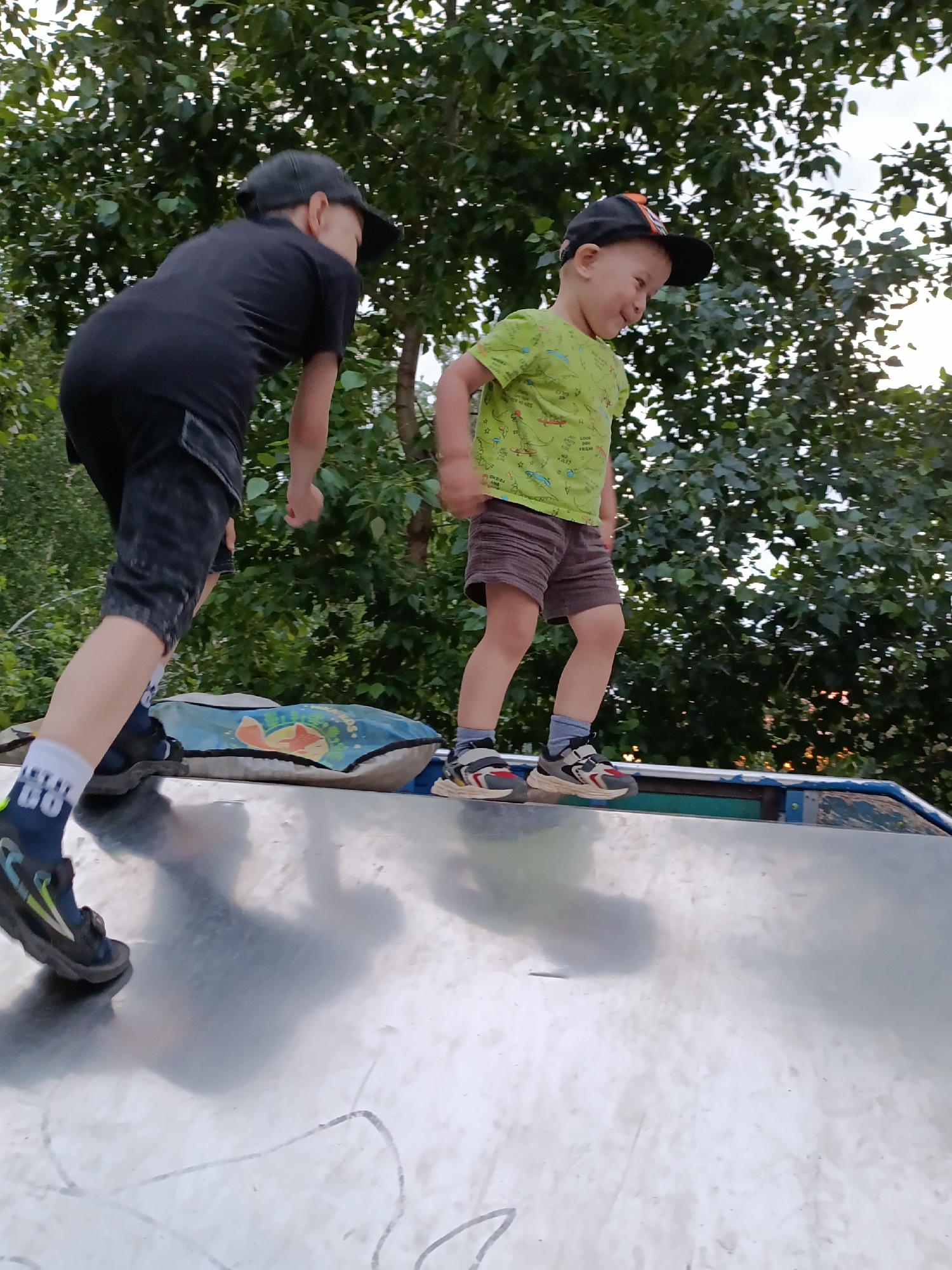 Two young boys are on a silver-colored slide in an outdoor setting, with trees and foliage in the background. The boy on the left, wearing a black t-shirt, black shorts, a black baseball cap, and dark sneakers with blue and yellow accents, is positioned closer to the foreground and is facing away from the camera. He appears to be in motion, with his left leg extended. The boy on the right, wearing a green t-shirt with a dinosaur print, brown shorts, and gray and red sneakers, is facing towards the left of the frame and looking down with a smile. He is also in motion, with his right leg bent. A blue and white patterned cushion with cartoon images is partially visible on the slide behind the boy on the left. The lighting suggests it is daytime, possibly late afternoon or early evening, with some sunlight filtering through the leaves. This scene is likely located in a park or playground within the city of Chaikovsky, Russia, given the environmental context and the commonality of such play structures in urban recreational areas.