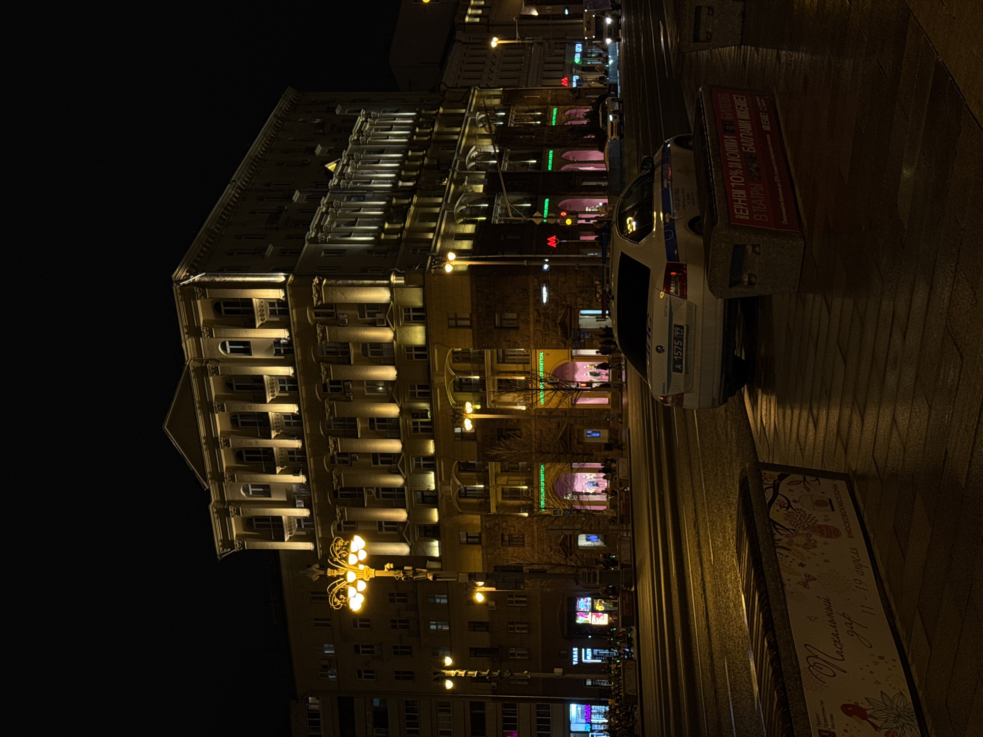The image depicts a night scene in Moscow, Russia. The focus is on a large, illuminated building with classical architectural elements such as columns and multiple windows. Street lighting, including ornate lampposts, casts a warm glow on the facade and surrounding area. A police car is parked on the side of the road, partially obscuring the view of the building. Signage for shops and metro stations (indicated by the "M" logo) is visible. A bench with advertising is placed at the roadside. Pedestrians can be seen in the background, suggesting a busy street. The image shows a blend of historic architecture and modern urban life.