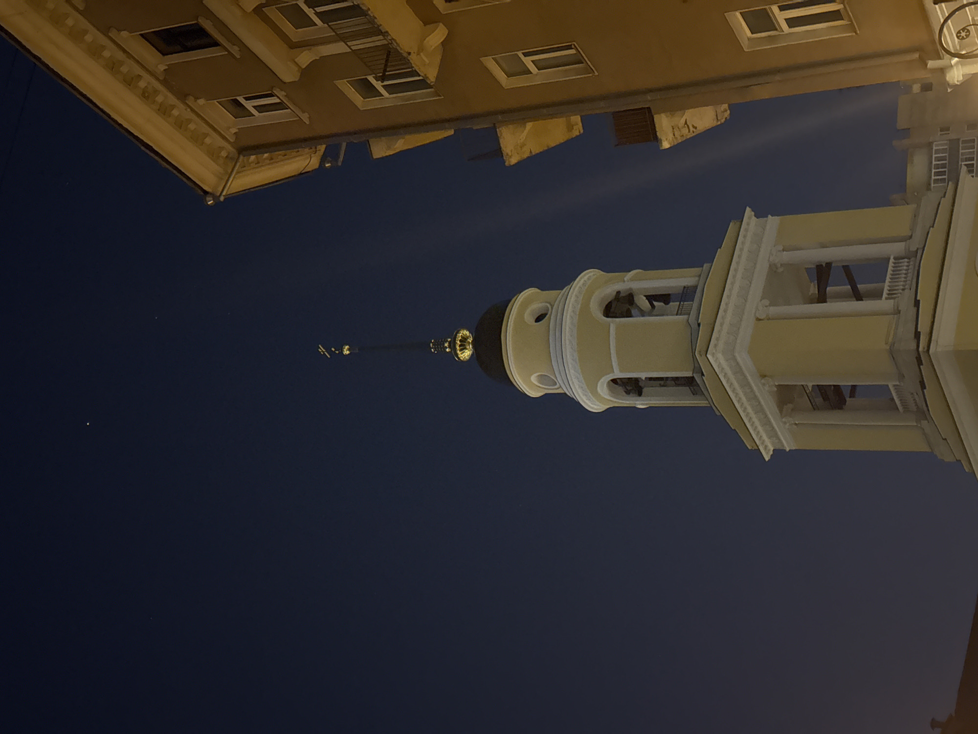 The image, oriented vertically, captures a striking night view of a historic architectural complex in Moscow, Russia, looking upwards towards a brightly lit bell tower. The central subject is a tall, slender bell tower, constructed from light yellow or cream-colored stone or stucco. It features multiple tiers, including a significant section with classical columns and arches, through which the interior structure or bells might be visible. The tower culminates in a dark dome, topped by a delicate spire and a prominent, gleaming golden ornament, possibly a cross or an orb, which reflects the light intensely.
Above and to the left, partially cropped, is another building of similar light color, showcasing windows and decorative cornices. This building appears to be part of the same complex or an adjacent structure.
The sky is a deep, dark blue, indicating either late dusk or nighttime. It is mostly clear, with a few very faint speckles of light, possibly distant stars or atmospheric phenomena, and one slightly brighter speck visible to the left of the spire. The architectural structures are illuminated by artificial lighting, which highlights their details and contrasts sharply with the dark sky. No people or visible text are present in the image. The perspective is from below, emphasizing the grandeur and height of the buildings against the night sky in Moscow, Russia.