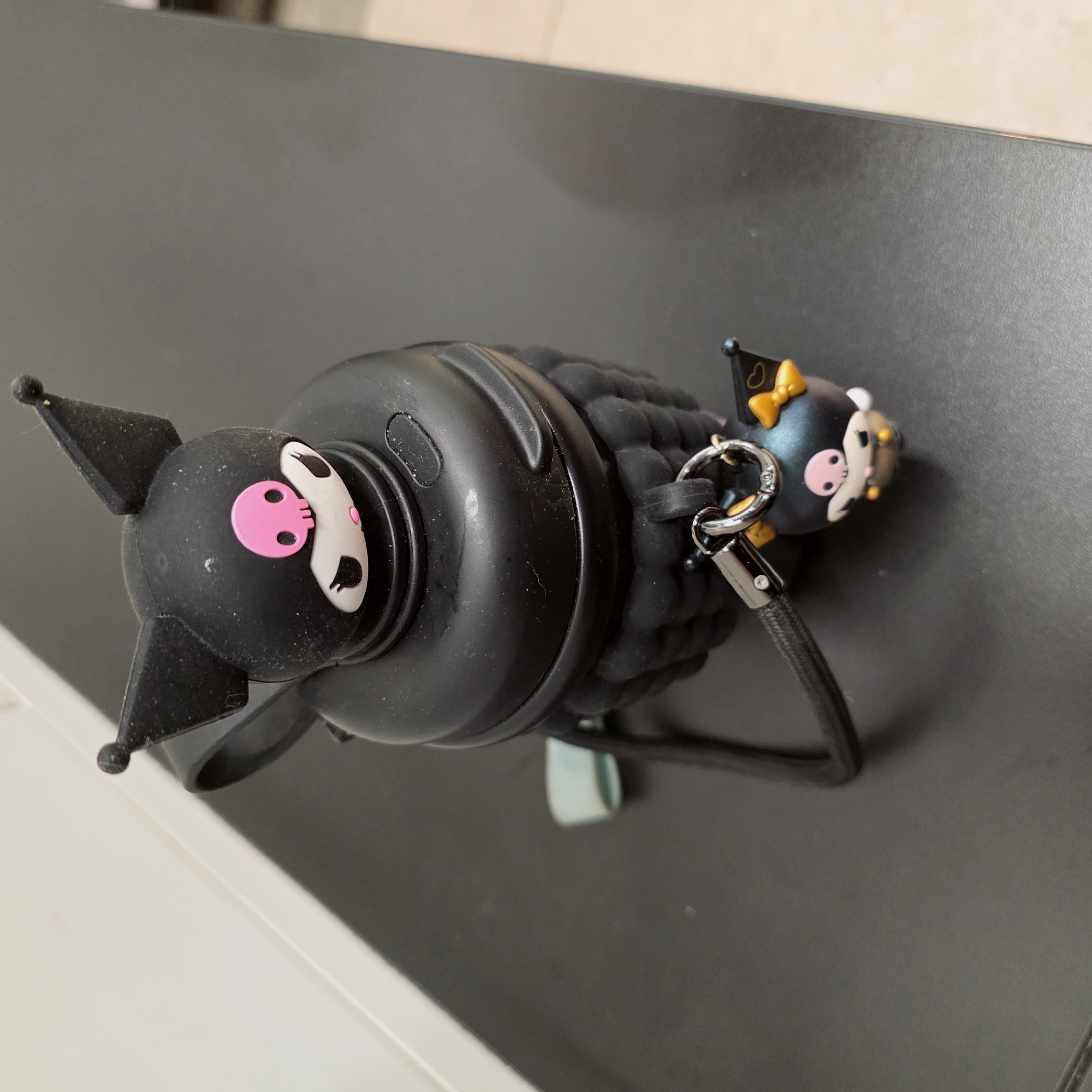 A black, cylindrical, character-themed object with a pink skull emblem is lying on a dark grey surface. A smaller matching character figure, also black, is attached via a ring and cord. The object's texture is bumpy and textured. The image was not taken in Saint Petersburg, Russia.