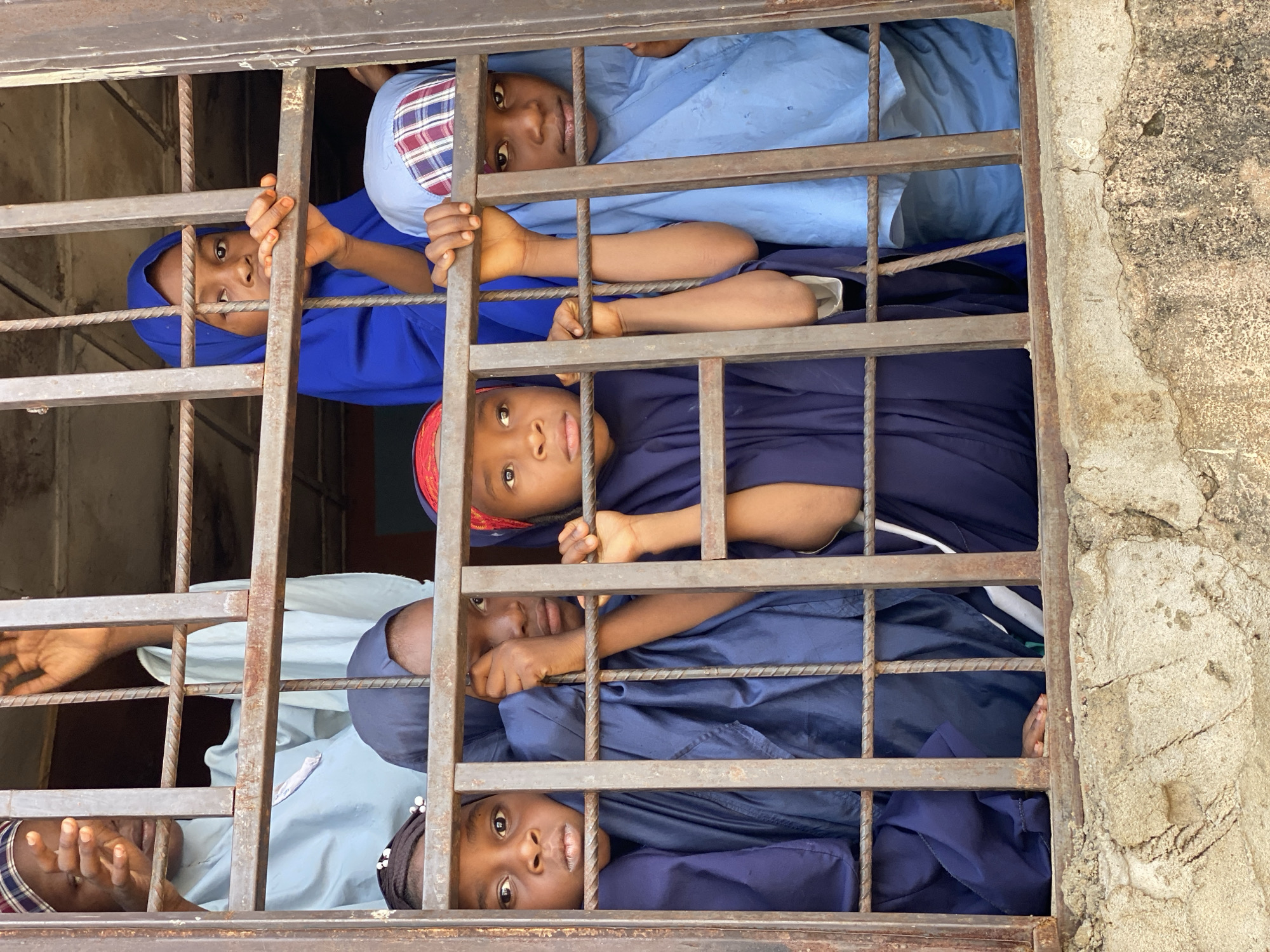 The image shows five young girls with dark skin peering out from behind a barred window. They are wearing what appear to be school uniforms with head coverings of different colors and patterns. The location is suggested to be in Sardauna, Nigeria, but specific environmental details are limited to the rusty metal bars of the window and a cracked wall to the right of the window frame. The girls are holding onto the bars, and their expressions suggest curiosity or hope.