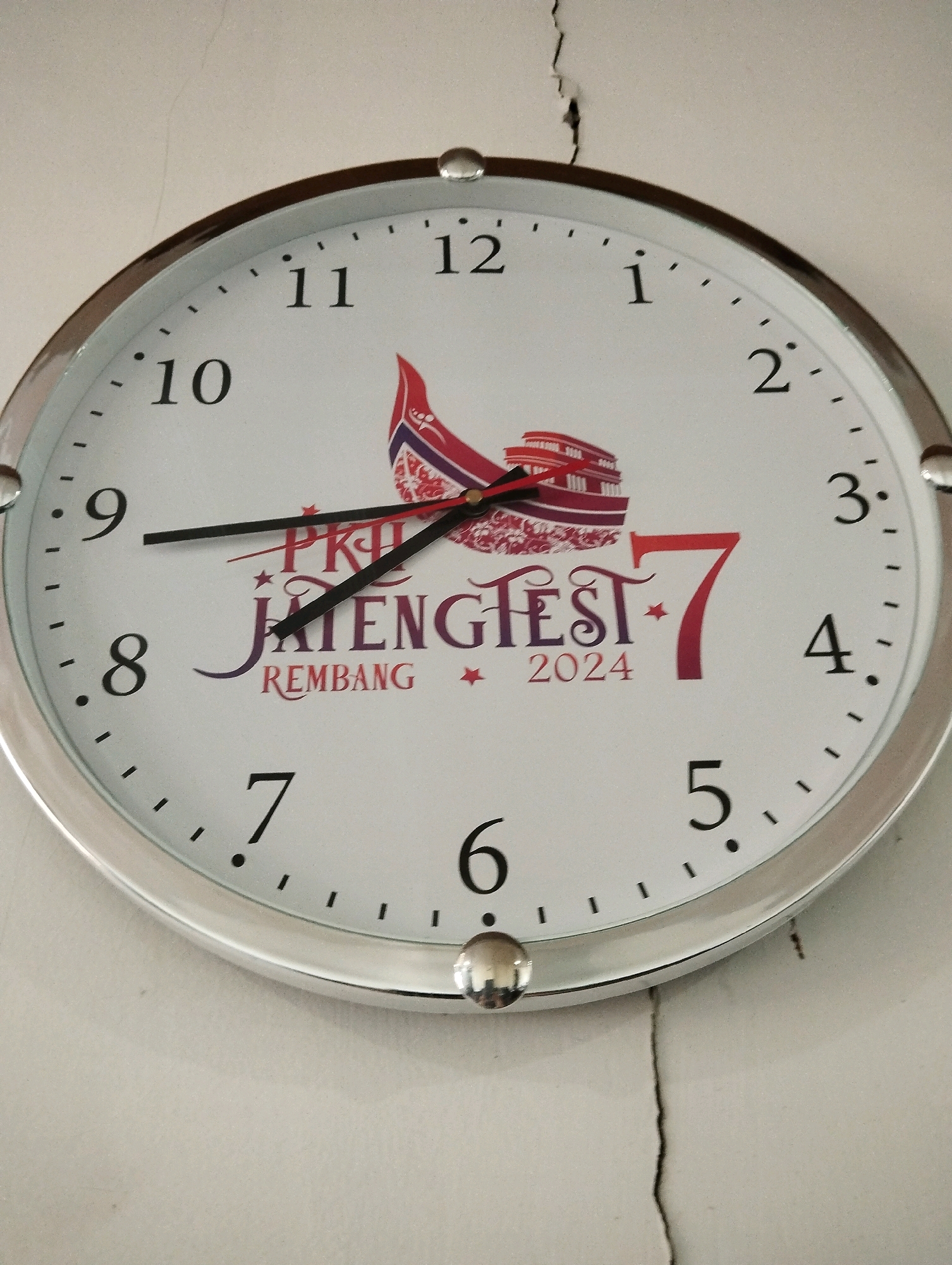 A close-up shot captures a white analog wall clock with a silver rim, mounted against a light-colored, textured wall exhibiting a prominent vertical crack. The clock face features black Arabic numerals for hours, ranging from 1 to 12. Red and black hands indicate the time. The clock also displays a logo with red and purple text that reads "PKJ JATENG FEST REMBANG 2024" and a stylized graphic of a building with a red roof. The hour hand is positioned between the 9 and 10, leaning closer to the 9, while the minute hand points slightly past the 10, indicating a time between 9:50 and 9:55. The image composition focuses on the clock itself, with the surrounding wall providing context for its placement. The specific location in Salatiga, Indonesia, cannot be determined from the visual information provided in the image.