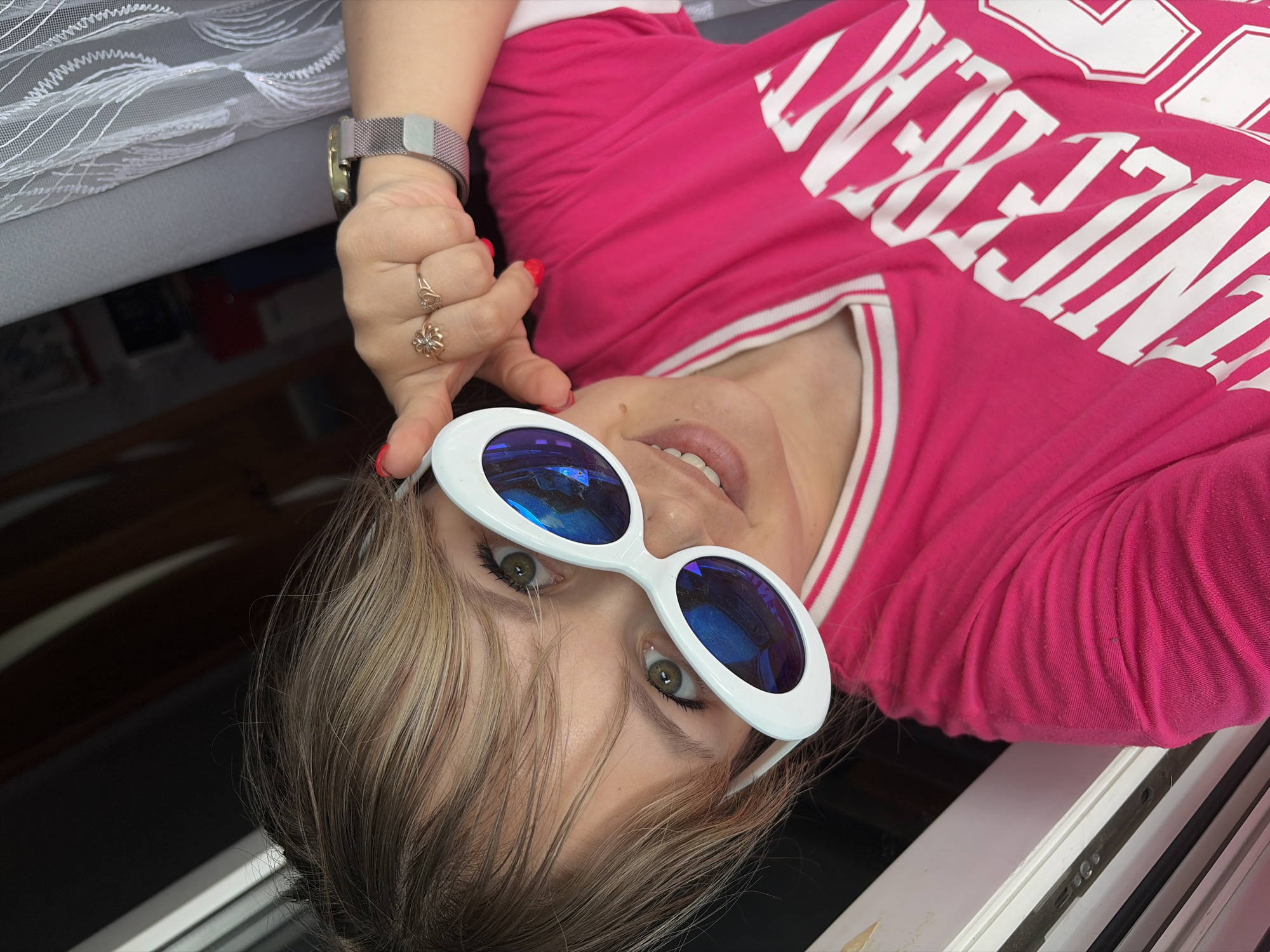 A person with blond hair and wearing white-rimmed sunglasses is looking at the camera. The person has a pink t-shirt with white lettering. They are also wearing a watch and rings. The background includes a white curtain and what appears to be part of a window frame. There is no indication that this image was taken in the city of Chaikovsky, Russia.