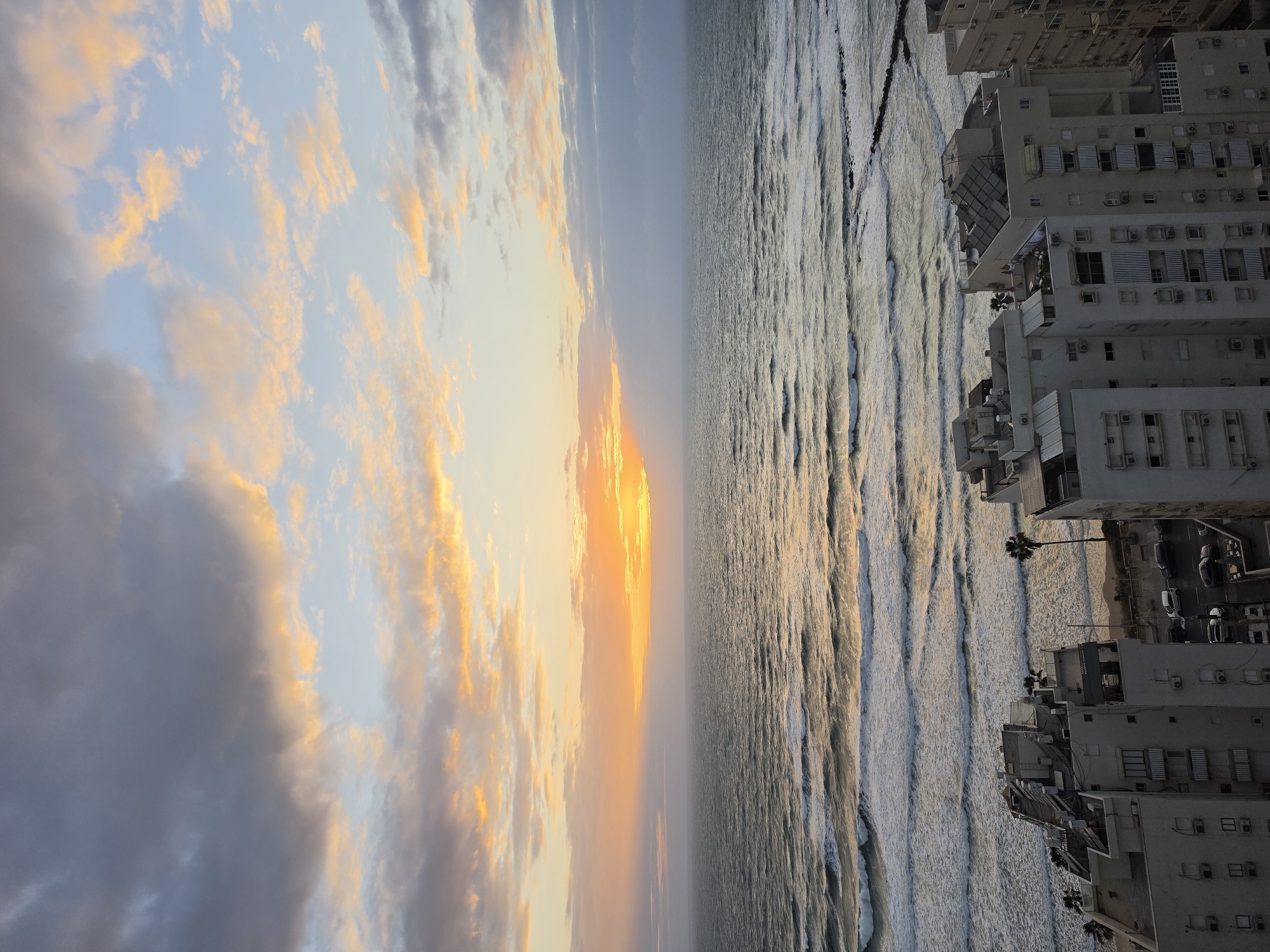 The image shows a scene in Bat Yam, Israel. The right side of the image depicts the coastline where buildings face the sea. The waves are visible as they come toward the shore. On the left side of the image, the sky shows a sunset with a blend of colors. The image is oriented vertically.