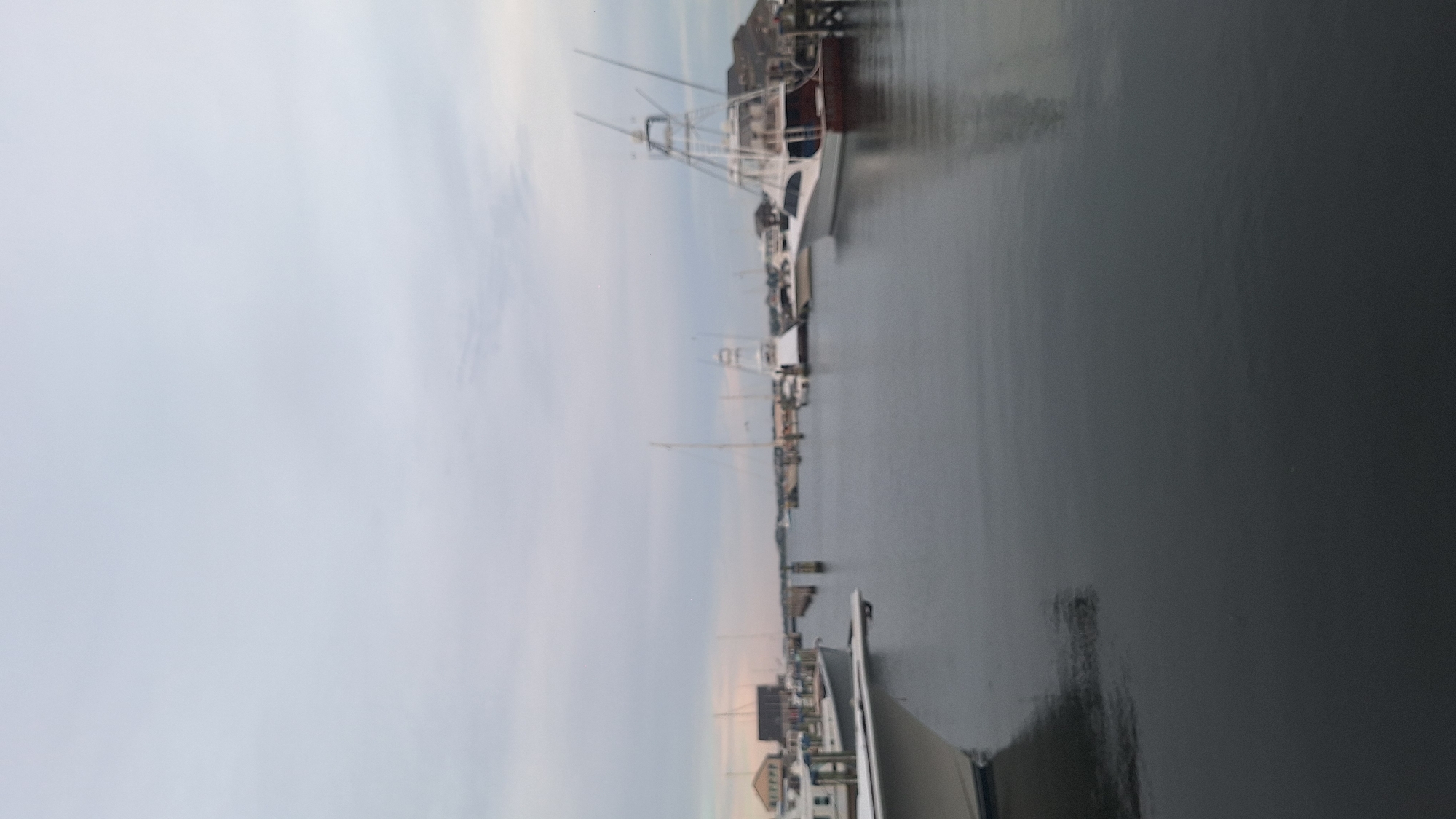 The image shows a harbor in Nantucket, Massachusetts, USA, with several boats docked in the water. The sky is overcast and the water is calm. In the foreground, a portion of a white boat is visible. In the mid-ground, there are several other boats, including a large white yacht with a tall tower. In the background, there are buildings on the shore and more boats. The overall scene suggests a quiet harbor, possibly in the morning or evening.