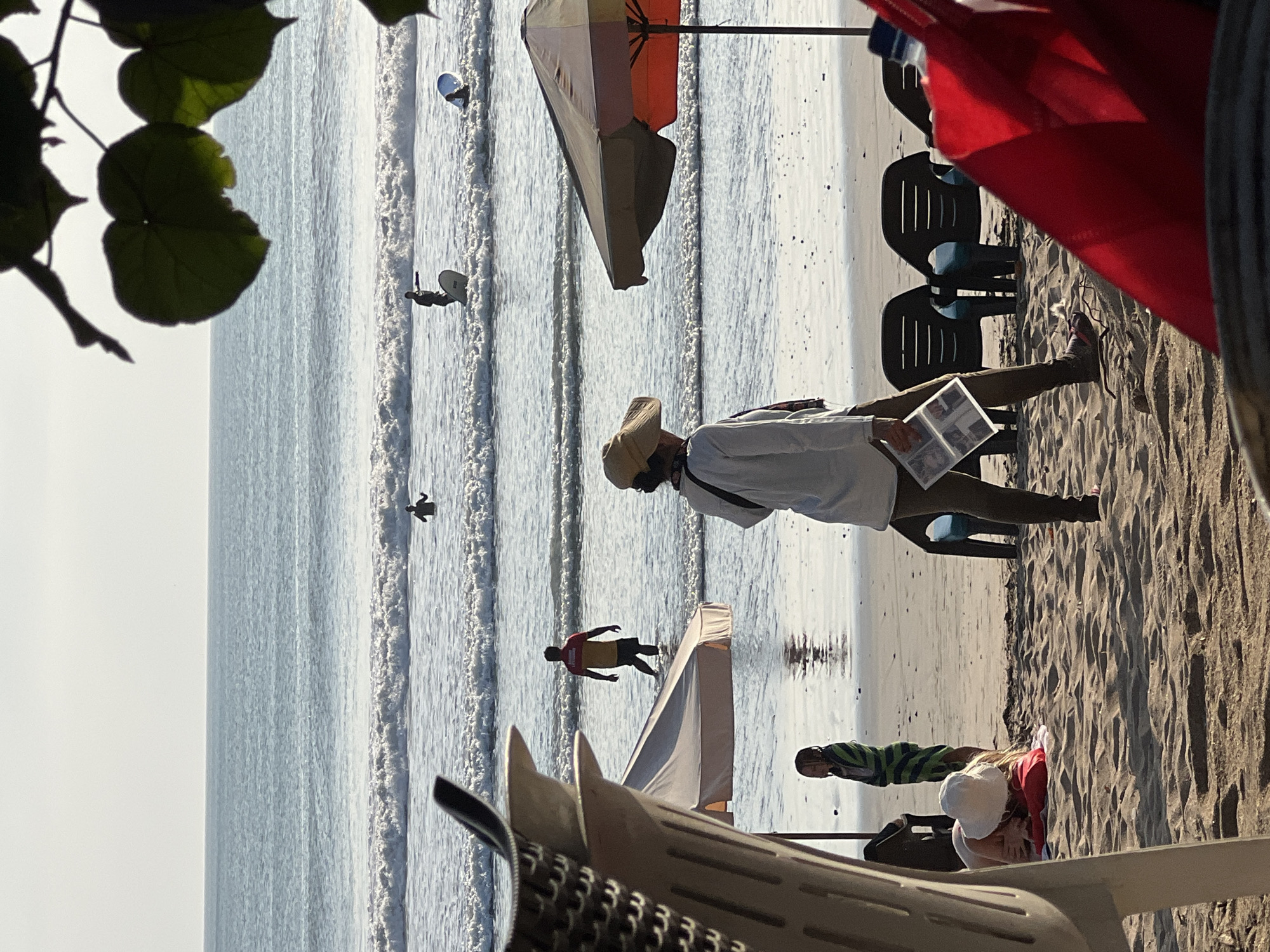 The image depicts a beach scene in Kuta, Indonesia, bathed in the warm glow of sunlight. In the foreground, a person wearing a broad-brimmed hat and a white shirt is walking away from the viewer, holding what appears to be a magazine or booklet. To their left, a child wrapped in a green and blue striped towel stands on the sandy shore. Further in the background, several beach chairs are arranged on the sand. The ocean stretches out to the horizon with gentle waves rolling in. There are several individuals in the water, some appearing to be surfers, with at least two on surfboards. One person is standing in the water near the shore, wearing a red and yellow shirt. The overall atmosphere is one of a relaxed beach day.