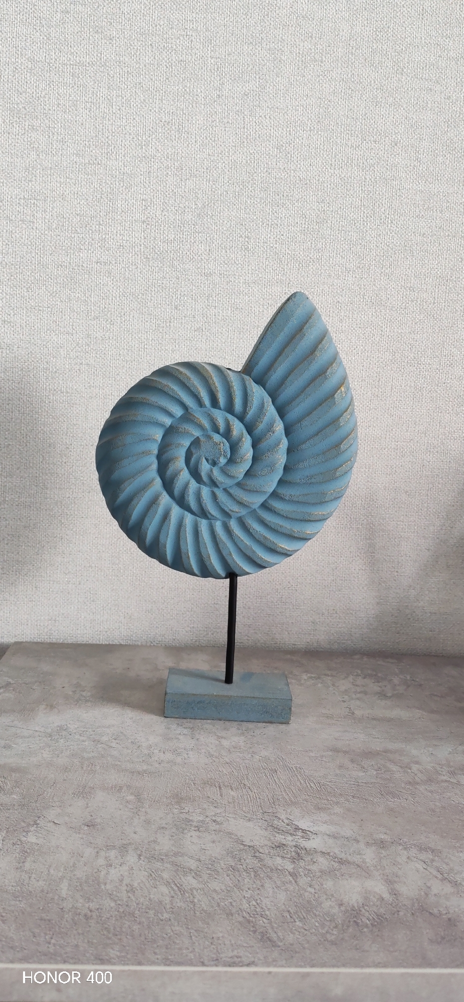 A nautilus shell sculpture painted blue rests on a stand against a plain, textured wall. The sculpture consists of a spiral shell on a black metal rod attached to a blue rectangular base. The surface beneath the sculpture is gray. The bottom of the frame bears the inscription "HONOR 400." No humans are present. It is not possible to tell the location from the visual data available.