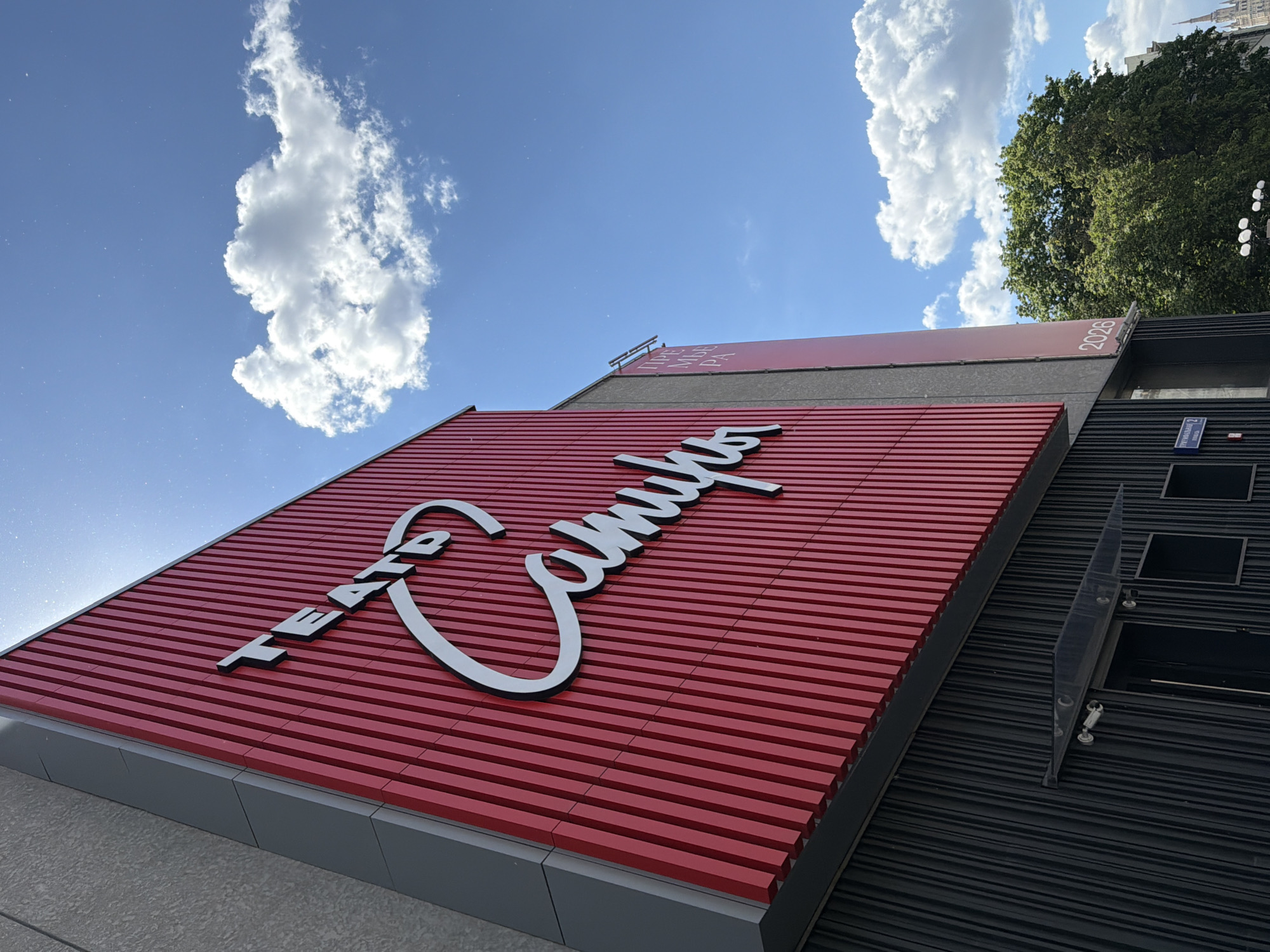 The image presents a worm's-eye view of the exterior of a modern building, likely a theater or cultural center, under a bright blue sky dotted with prominent white cumulus clouds. The building's design features distinct sections of corrugated panels.
The main focal point is a large section of the facade covered in horizontal red corrugated panels. On this red surface, a prominent white sign with a black outline displays the words "TEATRO Campu." "TEATRO" is oriented diagonally upwards to the left, while "Campu" is written in a more flowing, stylized script curving to the right.
Above this red section, a darker grey building element supports a red banner. On the far right of this banner, the numbers "2026" are visible. To the left, partially obscured white text in Cyrillic script can be deciphered as "ПРЕMЬEPA," which translates to "Premiere."
To the right of the red facade, the building continues with a section of dark grey or black horizontal corrugated panels. Attached to this darker wall is a small blue street sign with white text, clearly indicating "Гимназијска 2" (Gimnazijska 2) and "Нови Сад" (Novi Sad), placing the location in Novi Sad, Serbia.
The scene depicts a sunny day, with the building illuminated by direct sunlight. There are small bright specks, possibly dust or water droplets, visible against the clear blue sky in various parts of the image. A lush green tree and the faint outline of a distant spire are visible in the upper right background. No specific activity or people are shown, emphasizing the architecture and the bright, pleasant weather conditions.
