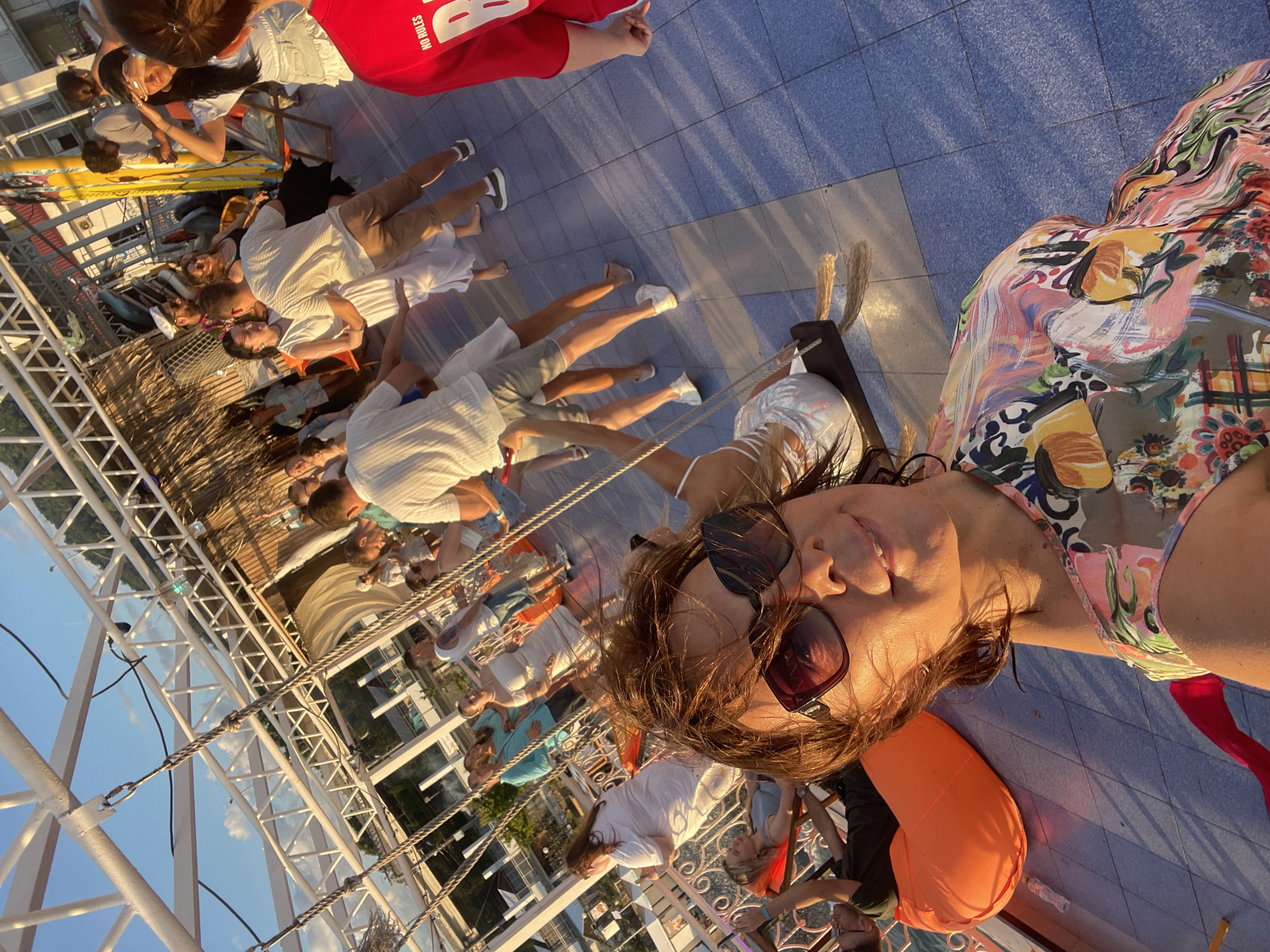The image depicts a crowded outdoor gathering, possibly a party or a festival, taking place on a raised platform with a blue tiled floor. The scene is bathed in warm, golden sunlight, suggesting it is either morning or late afternoon. In the foreground, a woman with sunglasses and a colorful patterned top is taking a selfie. She is positioned on a swing, with ropes visible above her. Behind her, a diverse group of people are engaged in various activities. Some are dancing, while others are conversing and socializing. One woman in a white dress is being lifted by a man, indicating a moment of celebration. The background reveals a backdrop of trees and buildings, hinting at an urban or semi-urban environment. There are also structural elements, such as metal beams and white supports, which might be part of a canopy or a larger construction. The overall atmosphere appears lively and festive.