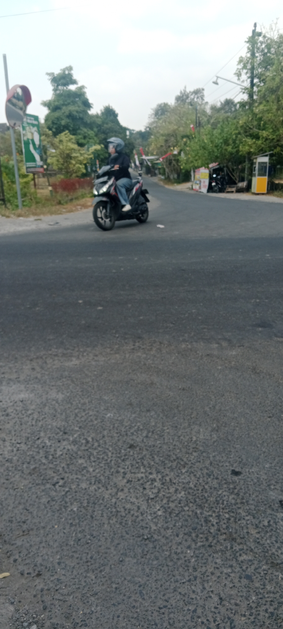 A person wearing a helmet and a dark jacket is riding a black motorcycle on a paved road. The motorcycle has a red and white design on its side. The person is wearing blue jeans and white shoes. The road appears to be in a rural or suburban area, with trees and vegetation visible on the sides. In the background, there are some buildings, including a small kiosk or booth with a yellow and white facade, and several parked motorcycles. A red and white barrier, possibly for traffic control, is also visible. A traffic mirror is mounted on a pole on the left side of the image. The sky is overcast. The location is identified as Maguwoharjo, Indonesia.
