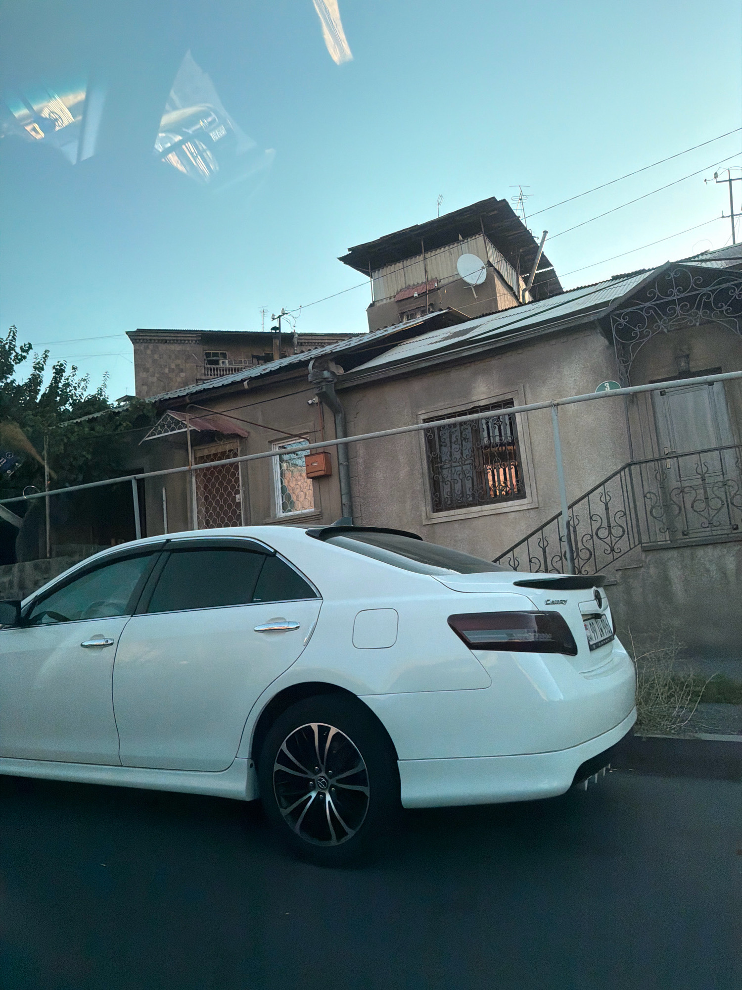 The image shows the rear and side of a white Toyota Camry sedan parked on a dark asphalt surface. The car's license plate is partially visible and appears to be Armenian. In the background, there is a light-colored building with a metal fence in front of it. The building has a dark, corrugated metal roof and several windows, some with decorative metal grills. Above the building, electrical wires are strung across a clear sky. A satellite dish is mounted on the roof of an adjacent structure. The scene appears to be in a residential area of Yerevan, Armenia, as indicated by the style of the buildings and the Armenian license plate. The lighting suggests it is either early morning or late afternoon.