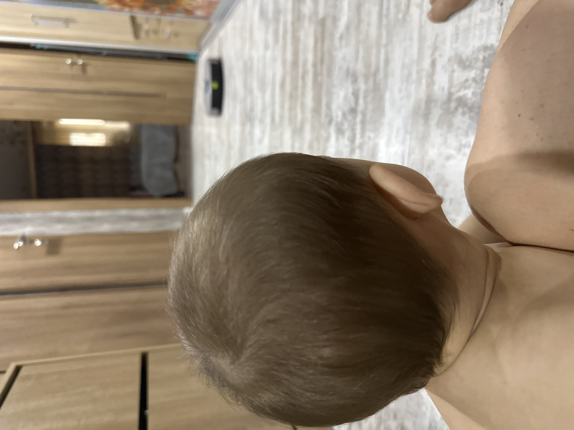 A close-up, rotated image features a baby's head and torso. The baby's light-brown hair is short and covers the scalp. The skin appears light-toned. In the background, blurred images of wooden drawers or cabinets, a cleaning robot and light-colored flooring can be seen.