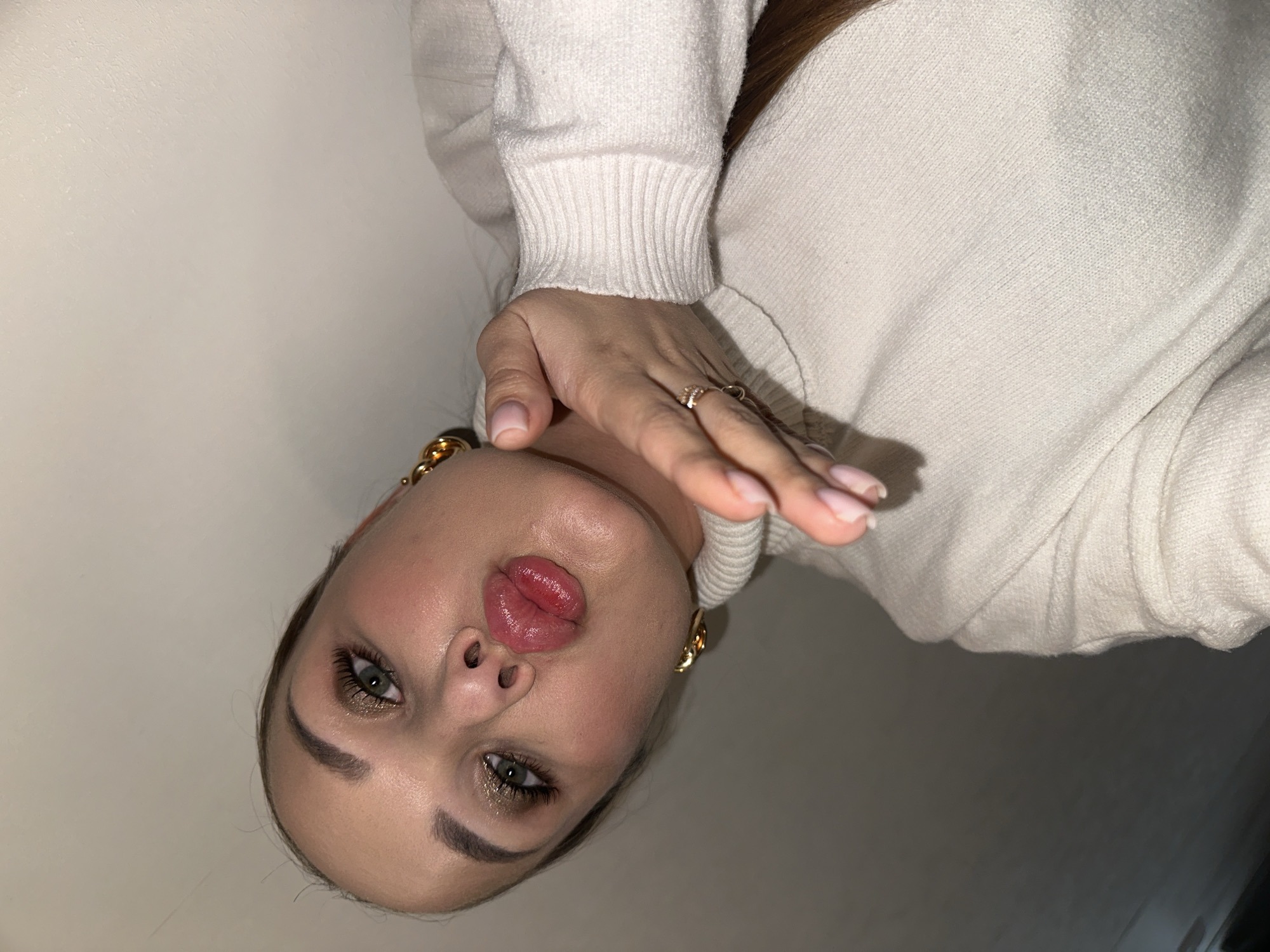 A young woman poses for a selfie. She is wearing a white sweater and gold hoop earrings. Her makeup includes eyeshadow and lip gloss. The background is a plain, light-colored wall. There is no information to confirm the location is in the city of Chaikovsky, Russia.