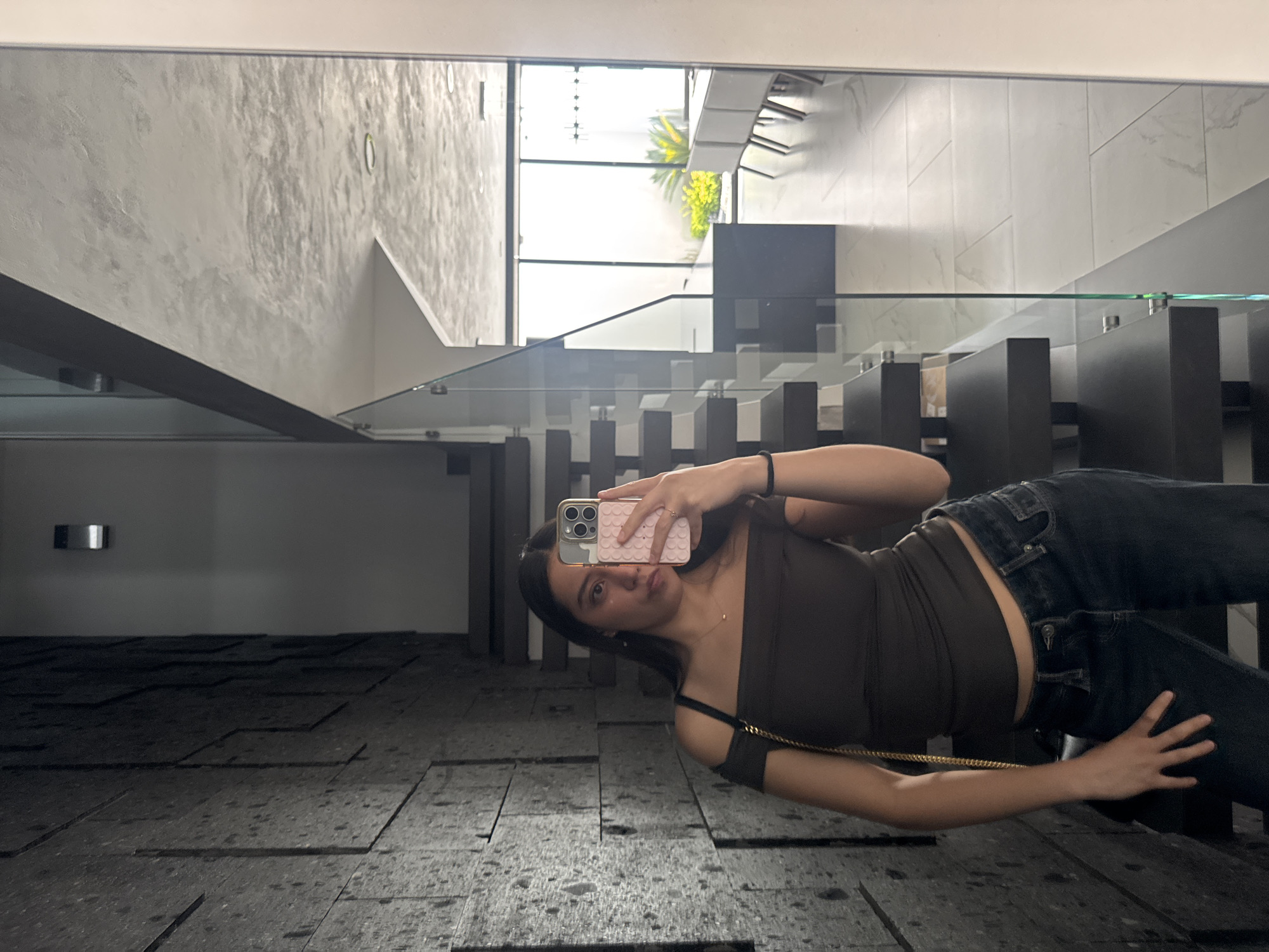 A young woman is taking a selfie in a modern building. The building has a glass staircase railing with black supports, and a large window overlooking greenery, possibly a palm tree. The interior walls are textured and light-colored, and the floor is dark with a tiled or paneled pattern. She is wearing a brown tank top and dark jeans. A gold chain bag strap is visible. The building appears to be well-lit and contemporary in design. It cannot be conclusively determined whether the location is in Puebla, Mexico based on this image.