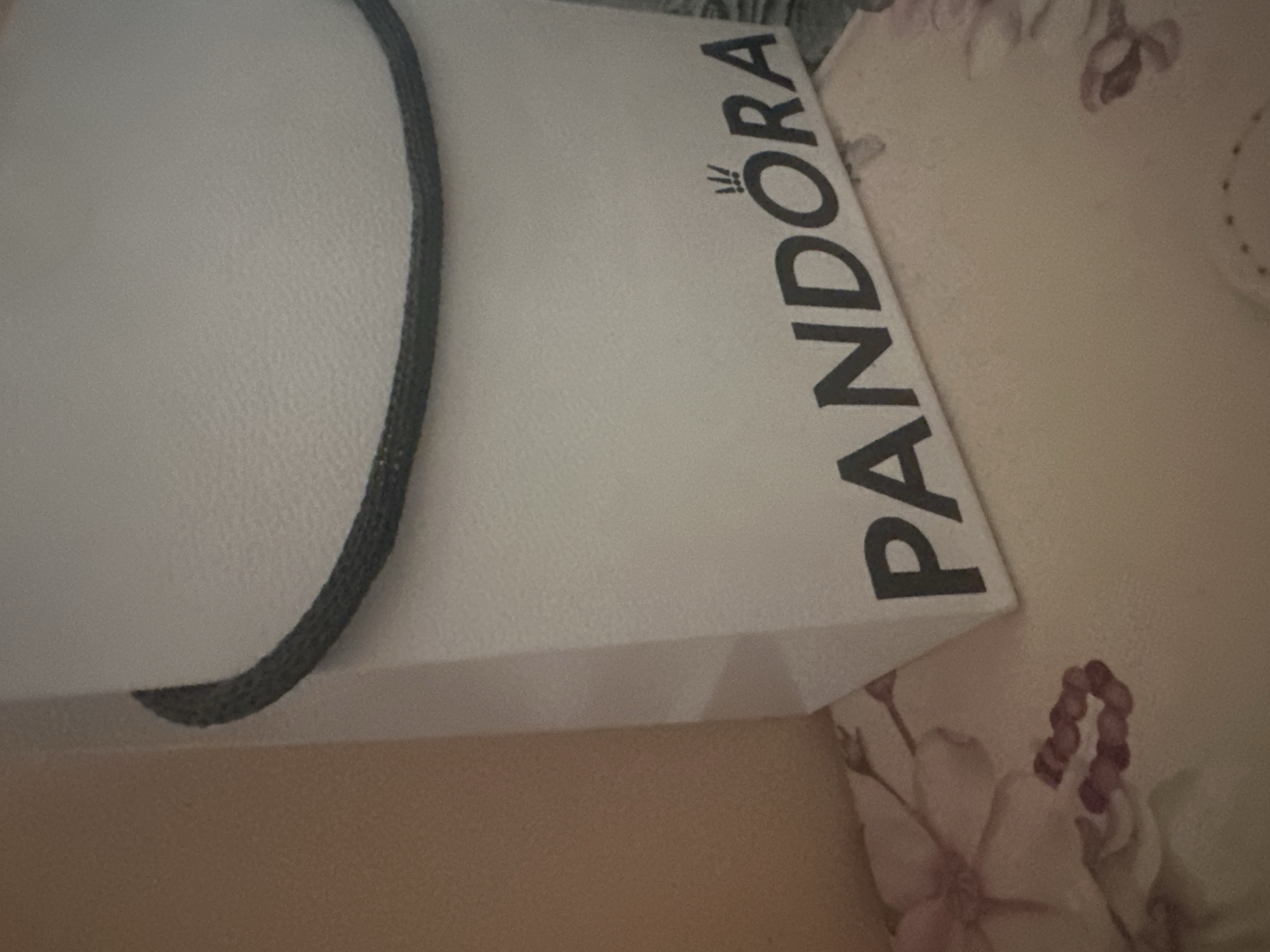 A close-up photograph shows a white Pandora shopping bag placed on a floral-patterned surface. The Pandora logo is printed in black on the bag. A gray strap handle is visible on the left side. The background shows parts of the floral design. The location and city of Sofia, Bulgaria, cannot be determined from this image.