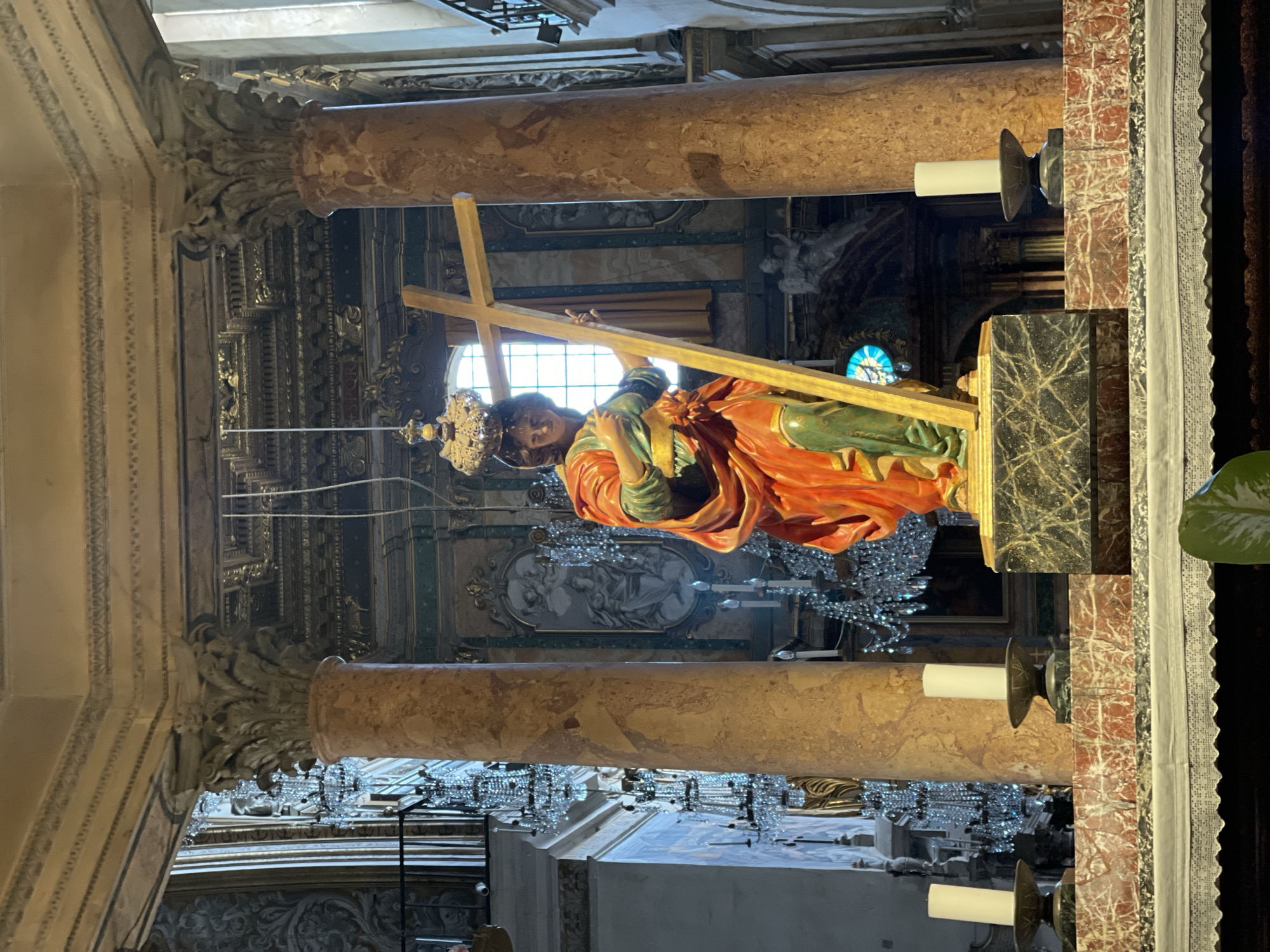 The image captures the interior of a church, likely in Rome, Italy, featuring a prominent statue of Jesus carrying the cross. The statue is placed on a marble platform and depicts a man in an orange robe and a green garment, wearing a crown and carrying a wooden cross. The background includes ornate architectural elements such as pillars, detailed carvings, chandeliers, and what appears to be a stained-glass window. There are candle holders on either side of the statue's platform. The overall scene evokes a sense of religious reverence and historical significance.