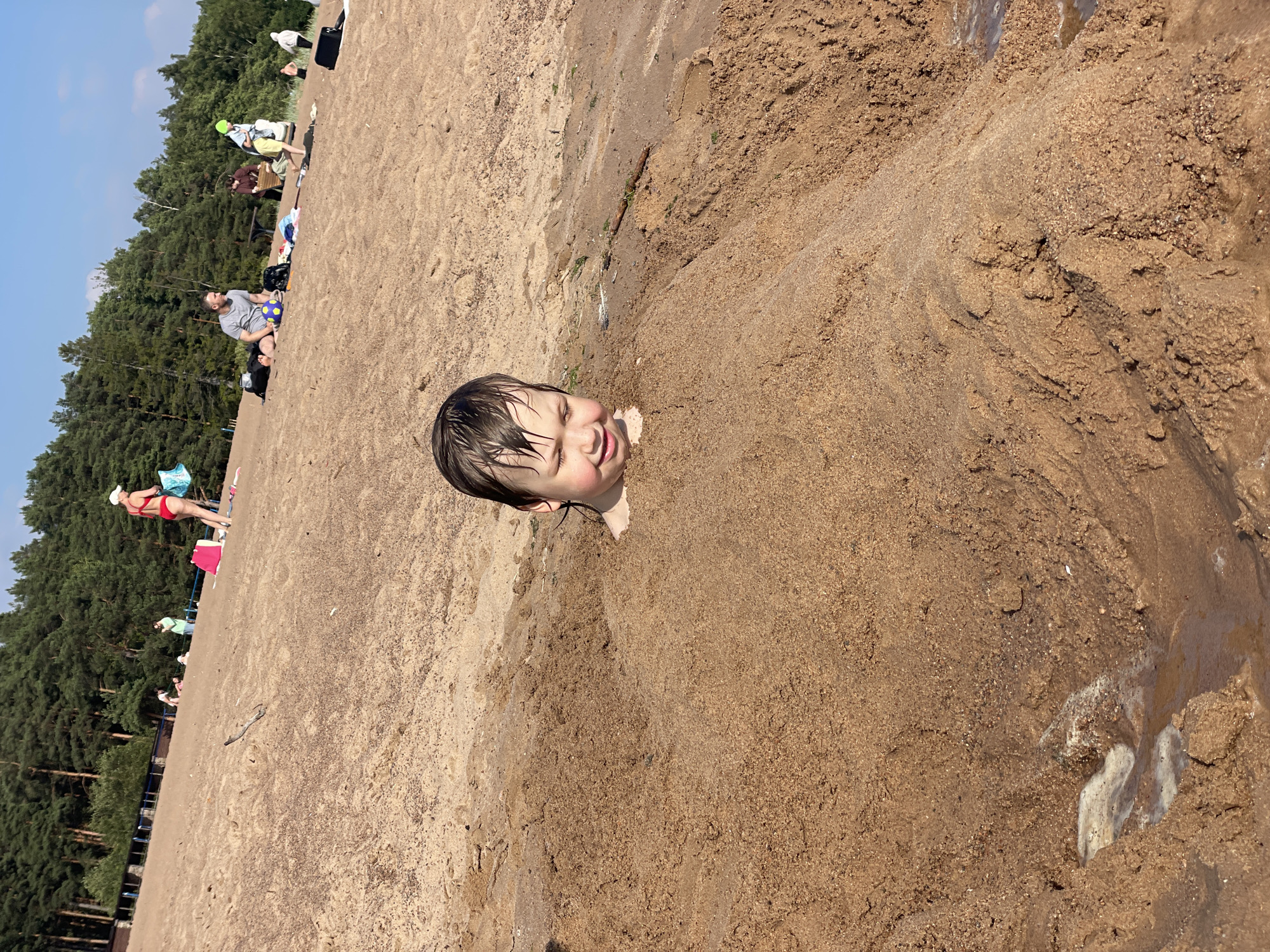 The image shows a young child buried in sand up to their neck on a sunny day at a beach. The child is smiling and appears to be enjoying the experience. In the background, there are other people on the beach, some sitting and relaxing, while others are walking. A line of trees forms the backdrop to the beach, suggesting a natural environment. The sand is damp in some areas, indicating it might be close to the water. The overall scene is one of a casual, leisurely day at the beach, likely during the summer given the attire of the people. The sun is bright, casting clear shadows, and the sky is blue with some white clouds. The location is identified as Solnechnoye, Russia. There is no visible text in the image.