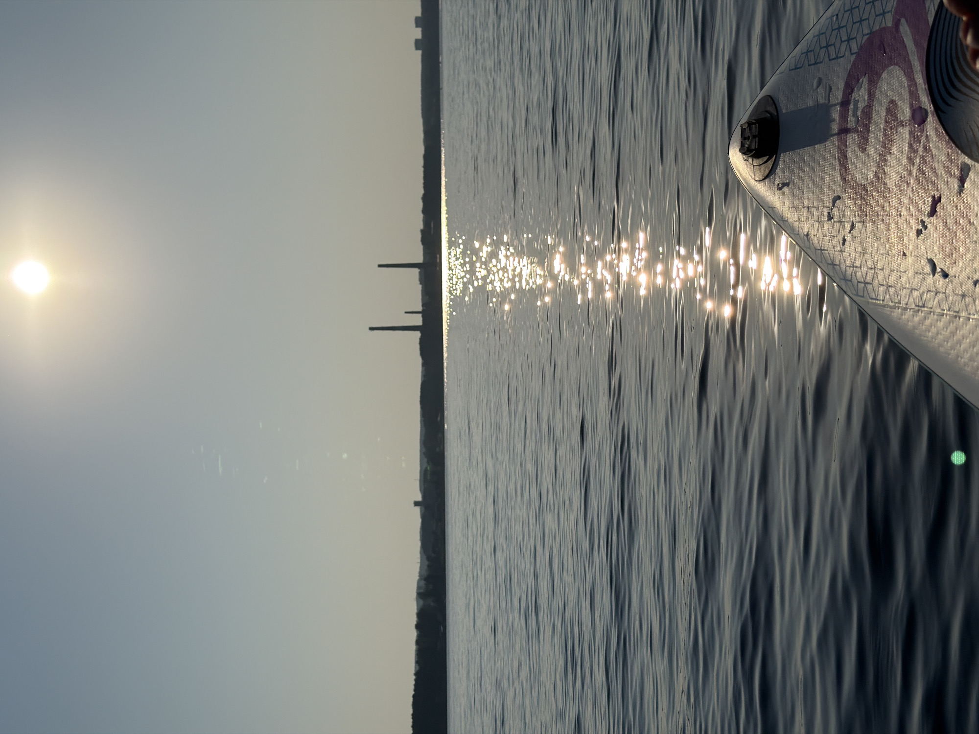 The image shows a view from a paddleboard on a body of water, likely the Kama River in Chaykovsky, Russia, given the presence of industrial smokestacks in the distance. The sun is high in the sky, casting a bright glare on the water and creating a shimmering reflection. The paddleboard's nose is visible in the foreground, showing part of its design and a black attachment. The water is calm with small ripples. In the background, a distant shoreline is visible, with trees and the silhouette of industrial structures, including tall, slender smokestacks, characteristic of a power plant or factory. The sky is clear and pale blue. The overall scene depicts a serene water activity with an industrial backdrop under a bright, sunny sky.