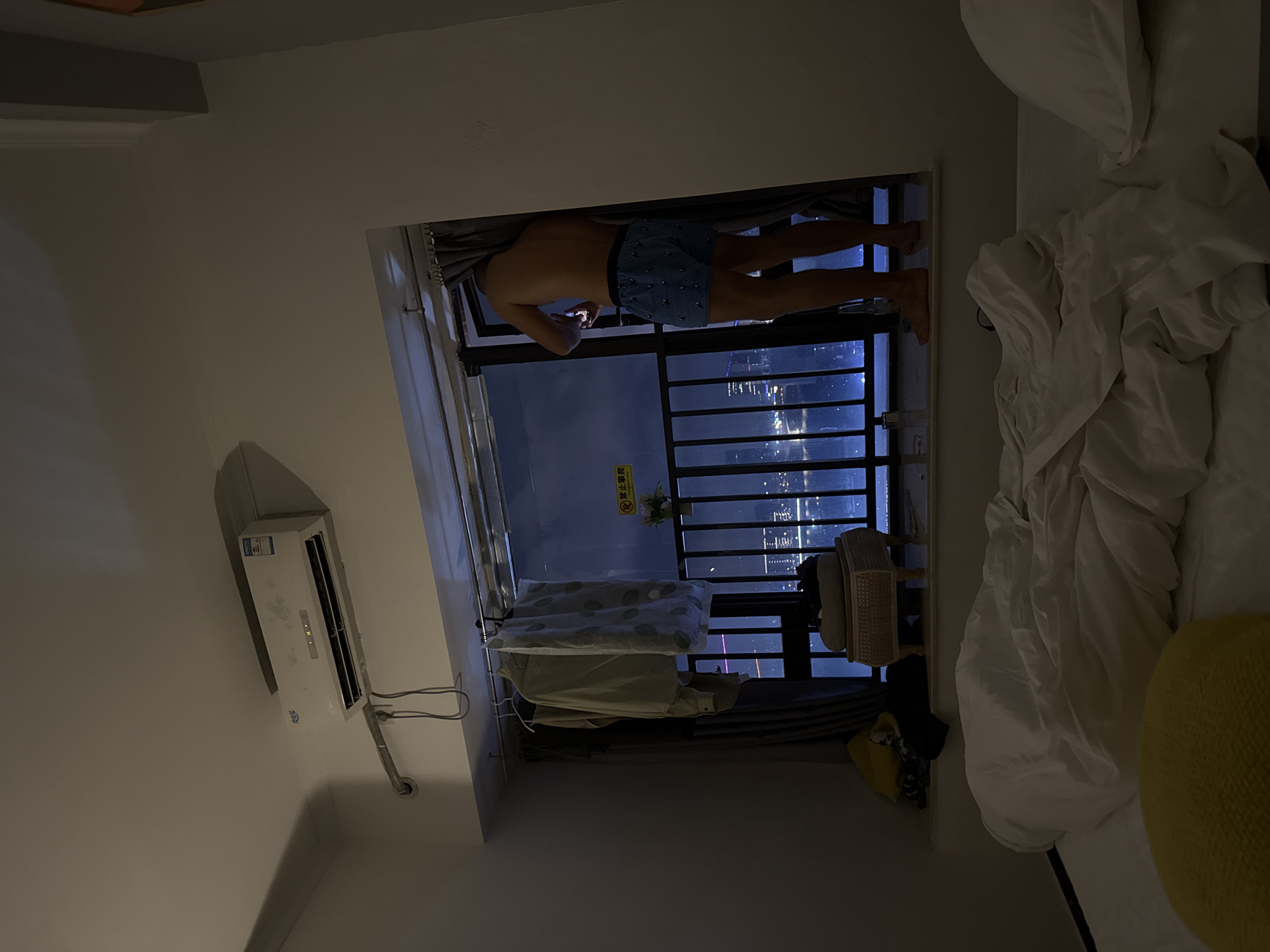 This image captures an indoor scene at night, likely a bedroom, with a view of a city skyline in Haizhu District, China. The room is dimly lit, with ambient light primarily originating from the exterior.
In the upper left, a white wall-mounted Midea brand (美的) air conditioner is installed, with a conduit and associated electrical wiring running beneath it. Spanning the top of the window frame is a metal rod from which several articles of clothing, including a light-colored shirt and a patterned fabric item, are suspended on hangers, appearing to air-dry.
The window features a dark metal safety railing. A yellow sign with black Chinese characters "禁止攀爬" (No Climbing) and smaller English text "NO CLIMBING" is affixed to the window glass or railing. A small potted plant with white flowers is placed on the window ledge near the railing.
A shirtless individual, wearing blue patterned boxer shorts, stands on the window ledge, facing away from the camera and observing the nighttime cityscape. Their bare feet are positioned on the ledge. The individual is holding a lit smartphone in their hands, appearing to interact with it. A dark curtain partially obscures the person on the right side of the window.
Beyond the railing, a dense urban environment at night is visible, characterized by numerous illuminated high-rise buildings and structures, with bright light reflections apparent on a body of water below.
In the immediate foreground, a bed with disheveled white sheets and a pillow occupies the lower portion of the frame. A yellow, textured circular cushion or pillow is partially visible on the bed in the lower-left. To the left of the person at the window, on an elevated ledge, a light-colored woven stool topped with a cushion is present, along with other dark fabric items.