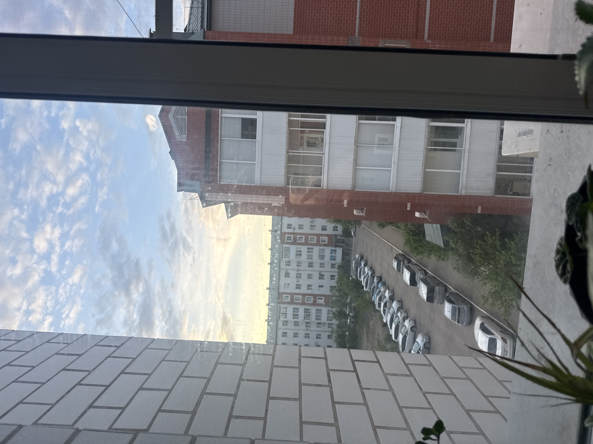 The image captures a view from a window overlooking a residential area, likely in Birobidzhan, Russia, given the architectural style of the buildings and the presence of parked cars. In the foreground, a white brick wall is visible on the left, and a window frame occupies the right side of the image, suggesting the vantage point is from inside a building.
Beyond the immediate foreground, a row of apartment buildings stretches across the middle ground. The building directly across from the viewpoint is a multi-story brick structure with numerous windows and balconies. To the left, a taller, light-colored apartment building with many windows is visible, indicating a densely populated urban environment.
A parking lot filled with cars is situated between the apartment buildings. Various makes and models of cars are parked in neat rows. The ground appears to be asphalt, and trees and greenery are interspersed around the parking area and along the sides of the buildings.
The sky above is partly cloudy, with a mix of white and grey clouds against a pale blue background. The lighting suggests it is either early morning or late afternoon, with a soft, diffused light illuminating the scene. The overall impression is one of a typical urban residential landscape. No people are visible in the image.