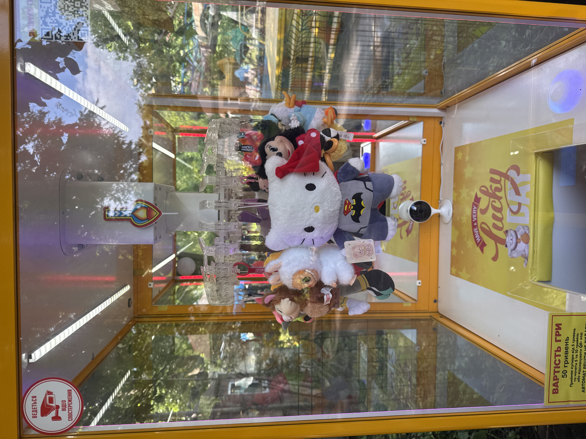The image shows a claw machine filled with plush toys, located outdoors in Vinnytsia, Ukraine. The machine has a bright yellow exterior with glass panels. Inside, various stuffed animals are suspended from the top by a mechanical claw. Prominent plush toys include Hello Kitty dressed as a superhero, Mickey Mouse, and a white bunny. The background suggests an outdoor amusement park or public space with trees and other attractions visible through the glass. A sign at the bottom of the machine indicates the cost of play, "VARTIST HRY 50 hryvnias" (cost of game 50 hryvnias), and specifies that it accepts 5 and 10 hryvnia coins or bills. There is also a circular sticker on the top left corner with a red border and a camera icon, reading "ВЕДЕТЬСЯ ВІДЕО СПОСТЕРЕЖЕННЯ" (Video surveillance is being conducted). A small security camera is also visible mounted on the inside bottom of the machine.