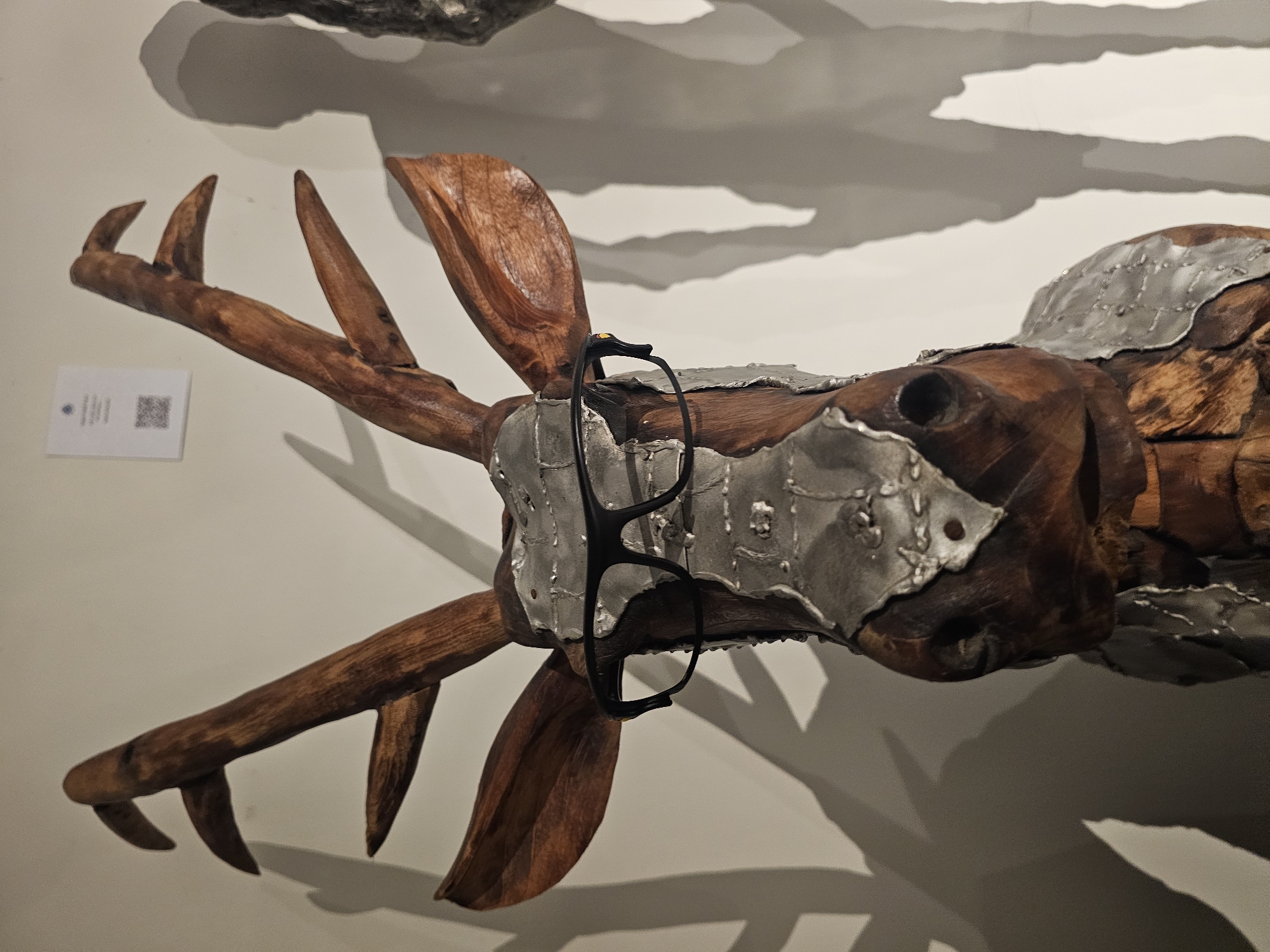 A close-up shot depicts a wooden sculpture of a deer's head adorned with silver metal plates and black-framed glasses. The sculpture is mounted on a white wall, casting shadows behind it. A small white label with a QR code is visible on the left side of the image. While the image does not provide explicit information about the sculpture's location within Denpasar, Indonesia, it appears to be an exhibit in a gallery or museum setting.