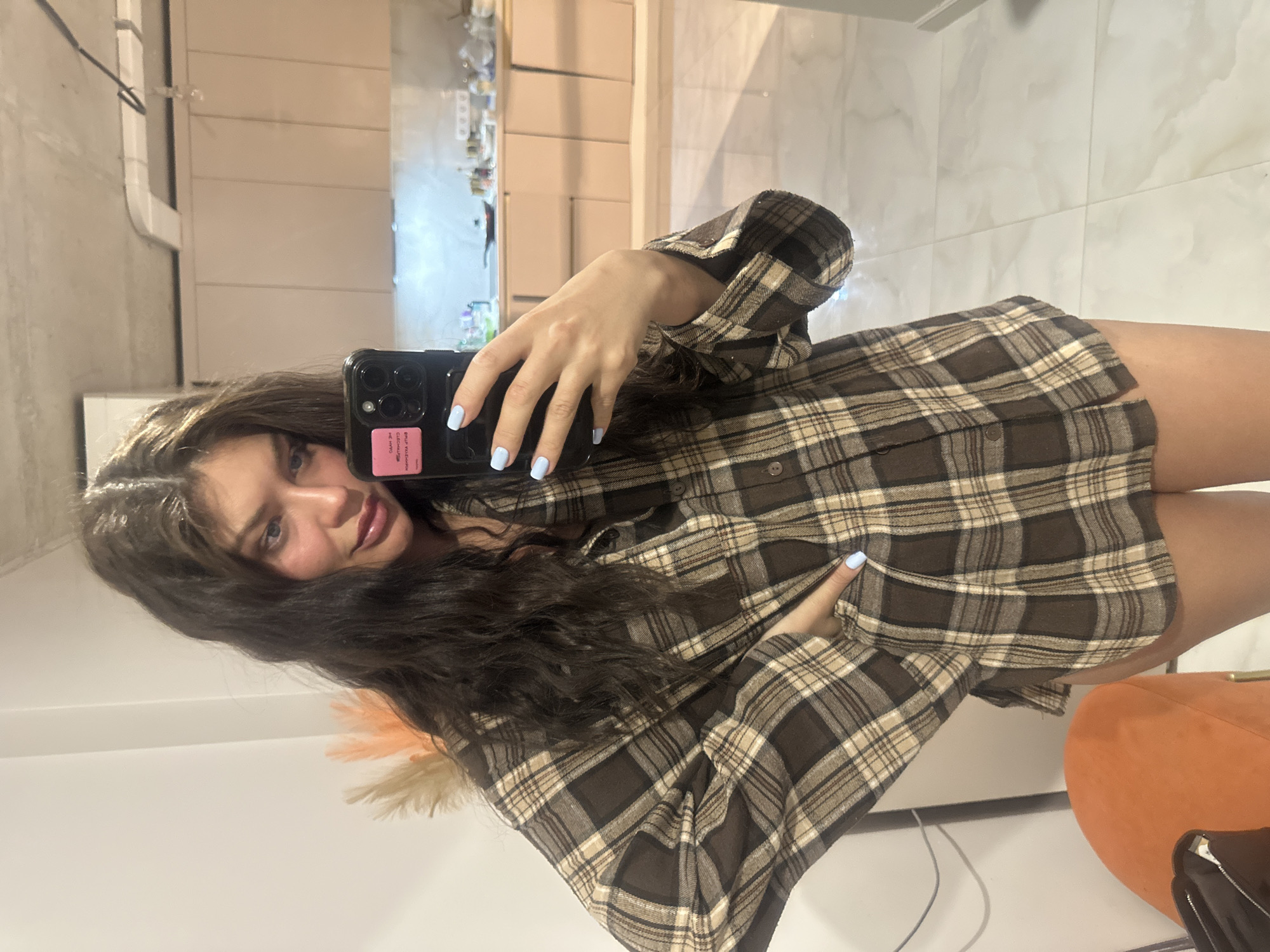 The image shows a young woman taking a selfie in what appears to be a modern apartment. She has long, wavy brown hair and is wearing an oversized, brown and beige plaid shirt. She is holding a smartphone with a pink sticker on the back, and her nails are painted a light blue color.
The background includes elements suggesting the location is inside an apartment. There is an orange, circular chair in the background. The location is not mentioned in the image, and therefore the exact location in Khimki, Russia cannot be determined.