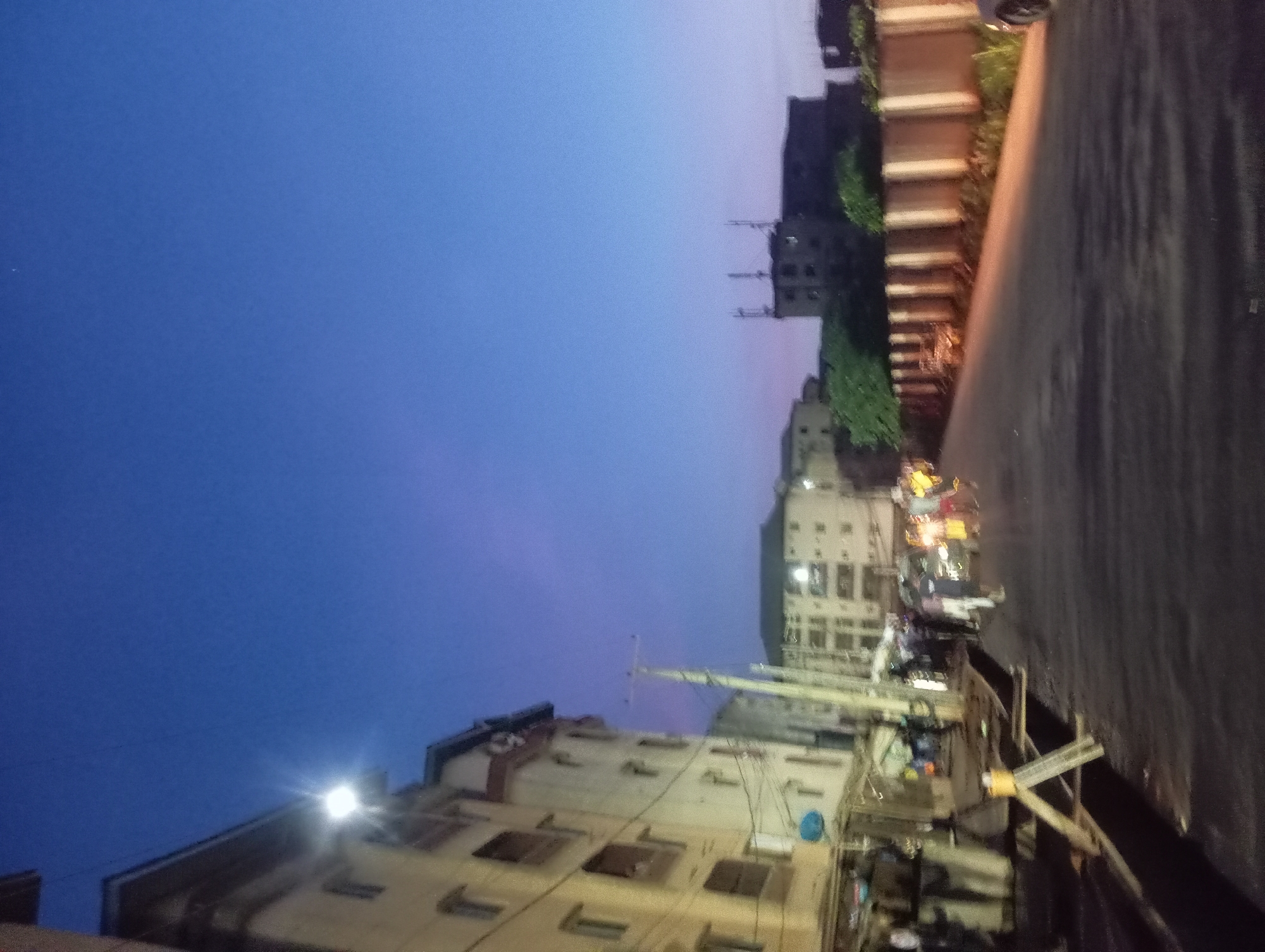 The image shows a street scene in Nkpor, Nigeria at dusk or dawn. A tall building with multiple windows stands on the left, illuminated by a bright light. Power lines are visible near the building. On the right is a road surface alongside a retaining wall covered with greenery. Further back, there are other buildings, some with visible antennas. Traffic is present on the street, with blurred lights suggesting vehicle movement. The sky is a gradient of blue and purple.