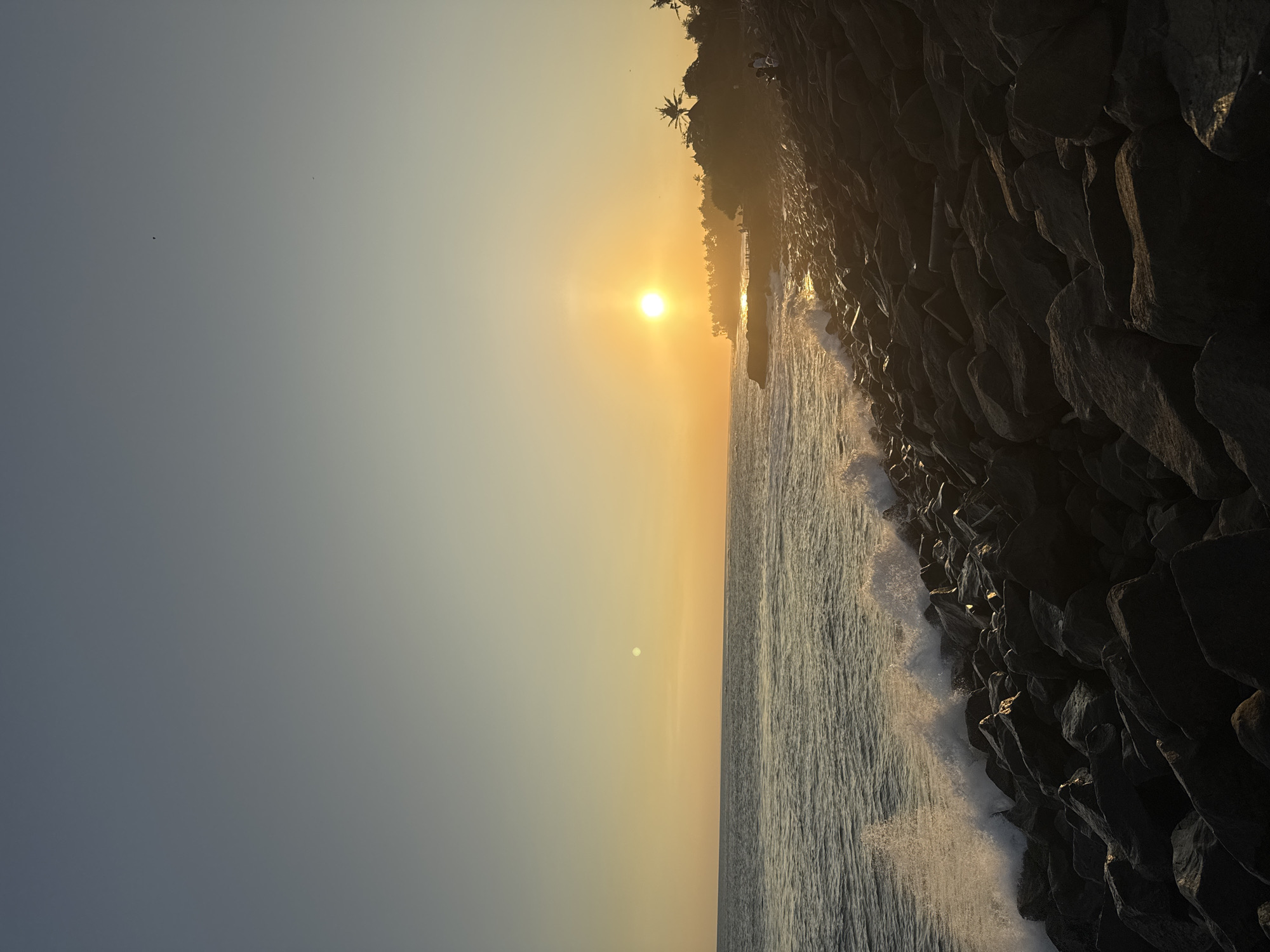 The image shows a shoreline at sunset near Mengwi, Indonesia. Large, dark, basalt rocks form a breakwater along the right side of the image, with ocean waves crashing against them. The sun is positioned low on the horizon, casting a golden glow across the sky and sea. The sky transitions from a blue-grey on the left to a yellow-orange near the sun. Silhouetted palm trees are visible on the land. The sea has a yellow tinge to it, that grows brighter and brighter, as the viewer approaches the sun, because of the reflection of it on the sea.