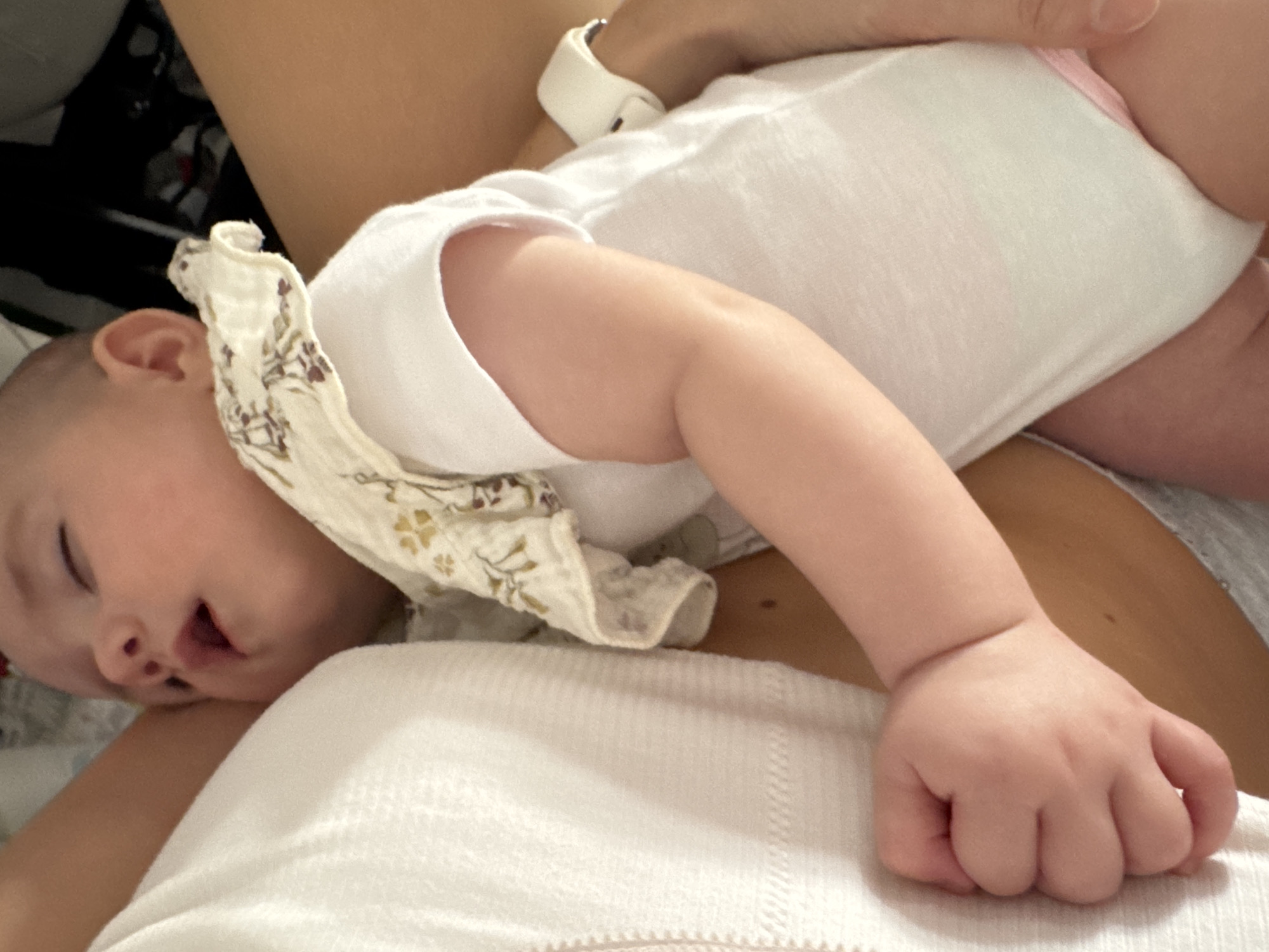 A baby is sleeping on its back, held close to another person's body. The baby is wearing a white onesie with a light-colored bib featuring a floral pattern around its neck. The baby's left arm is extended and resting on a white textured fabric. The other person's skin is visible, with a white watch on their wrist. The background is out of focus, showing dark shapes. The scene appears to be indoors, likely in a home environment, and does not contain any elements that would allow for its location to be determined as Isola delle Femmine, Italy.