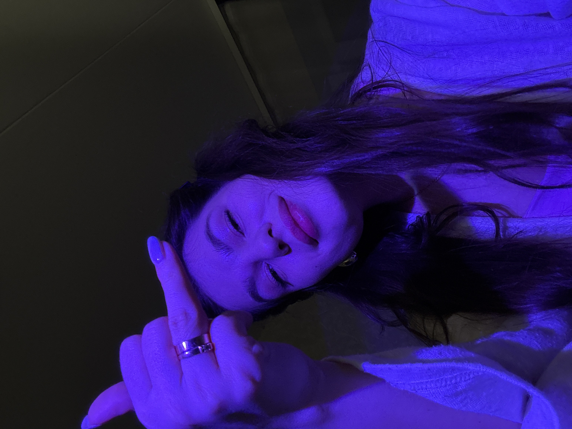 A woman with long, dark hair and fair skin is facing the camera, holding up her middle finger. Her nails are painted blue. She is wearing a gold ring on her finger and gold earrings. The lighting is a strong blue hue. The background appears to be a dark surface and light-colored fabric. There is no information in this image that indicates the location is in Chaikovsky, Russia.