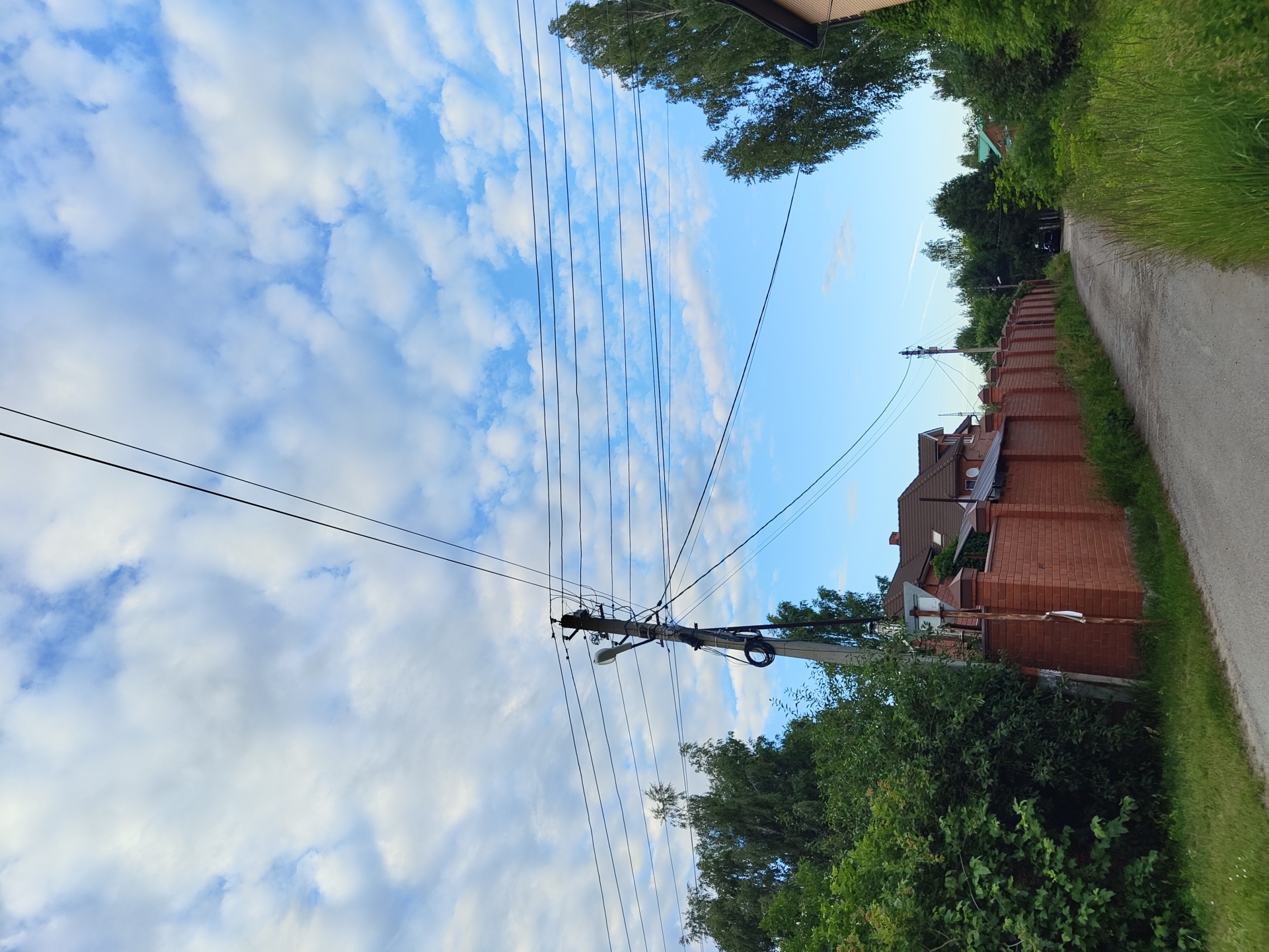 This media file depicts a roadside scene in Tsibino, Russia. The image is taken in a vertical orientation, capturing a road on the right side of the frame, bordered by lush green grass and trees. Overhead, a clear blue sky with scattered white clouds dominates the upper portion of the image. A power pole with multiple wires stretches across the frame, with a street lamp attached to it.
To the left of the road and behind a prominent brick wall, several houses with tiled roofs are visible, nestled amongst dense green foliage. The overall setting suggests a semi-rural or suburban environment. The time of day appears to be late afternoon or early evening, indicated by the soft lighting and the clear sky. There is no activity or event clearly taking place, other than the presence of a parked car in the distance down the road. No text is visible in the media file.