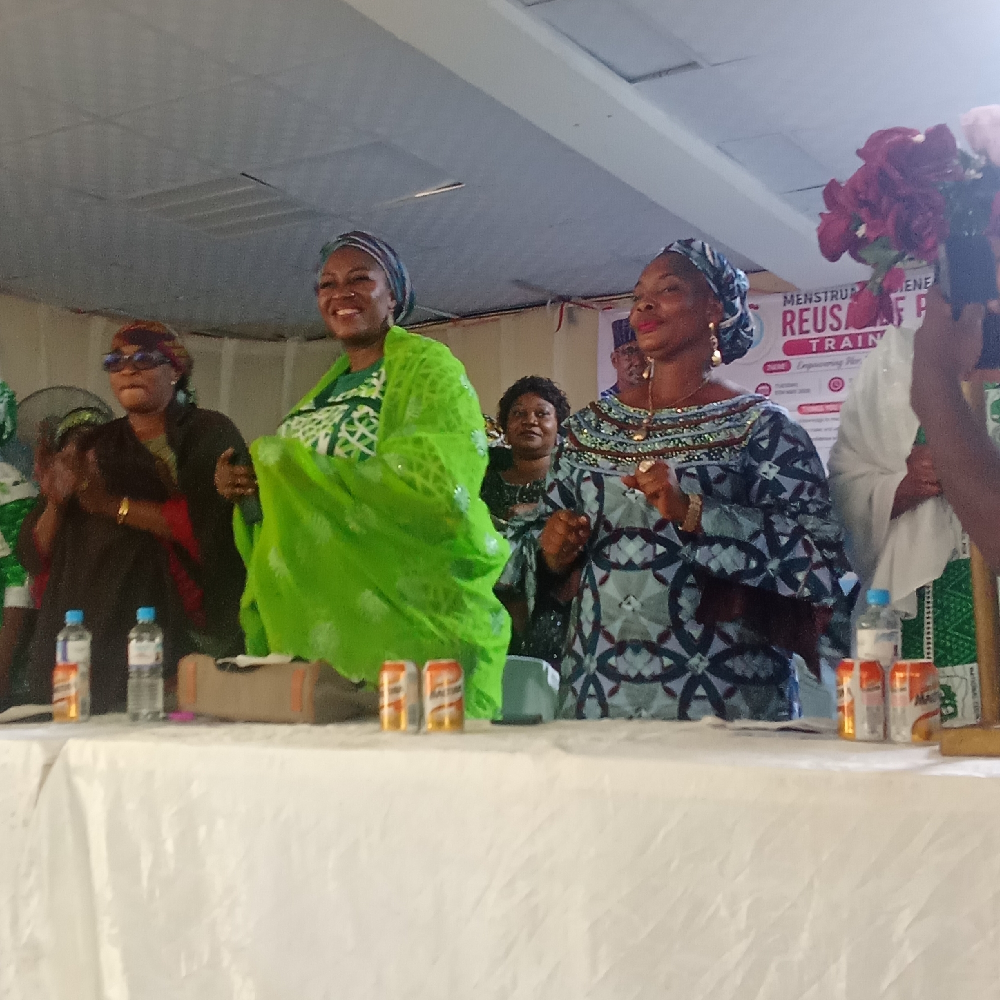 The image depicts a group of women at an event in Akwanga, Nigeria. Three women are prominently positioned behind a table covered with a white tablecloth. The woman on the left wears sunglasses and a dark outfit with red accents, while clapping. The woman in the center is dressed in a bright green outfit and appears to be dancing or moving dynamically. The woman on the right is wearing a patterned blue and grey outfit. In the background, other women are visible, as well as part of a banner with the words "MENSTRUAL HYGIENE" and "REUSABLE PADS TRAINING" visible. Water bottles and cans are placed on the table in front of the women. A hand holding flowers extends into the frame on the right. The room appears to be well-lit.