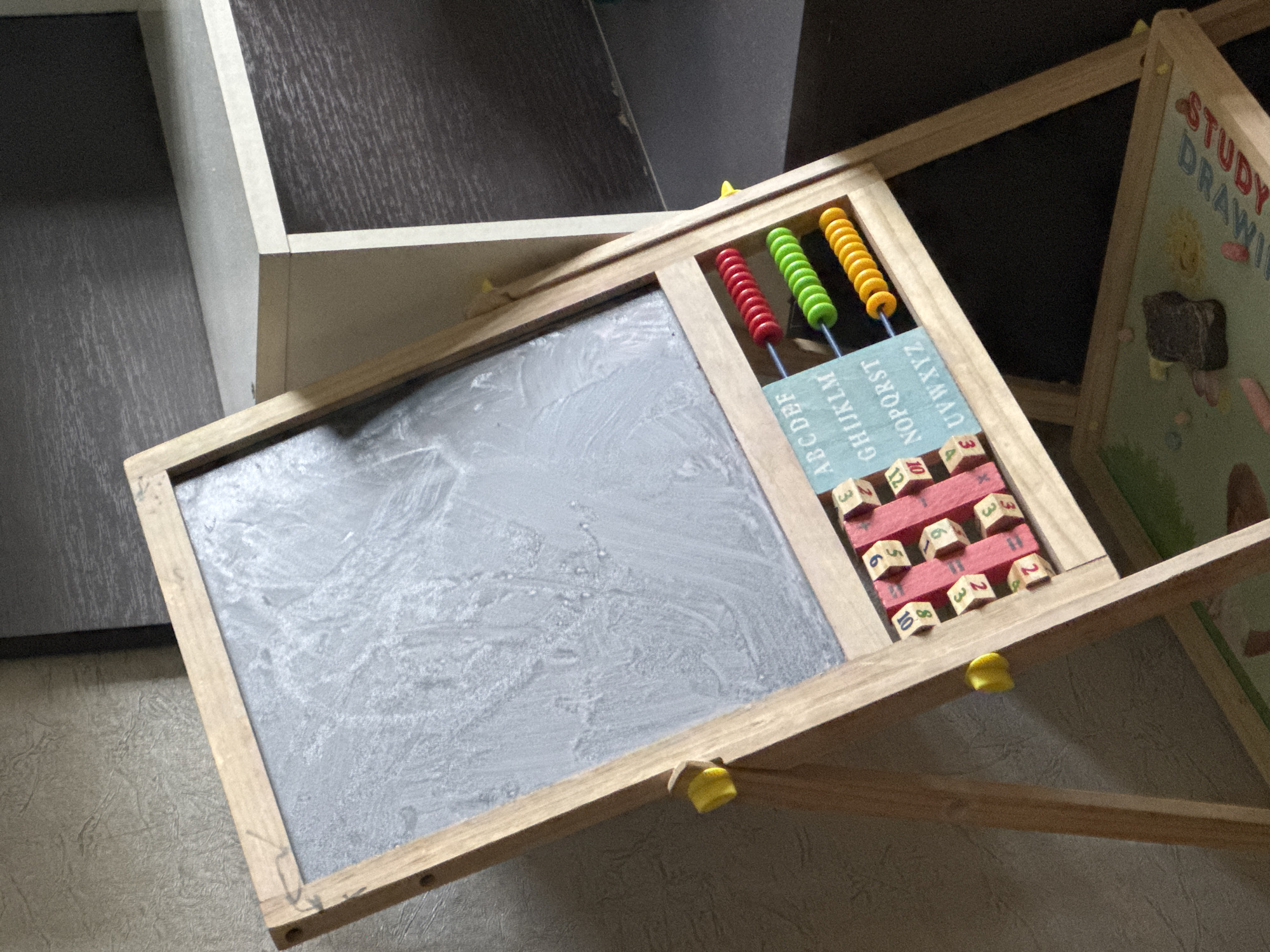 An easel-style children's chalkboard, made of wood, is set up indoors against a textured wall. The chalkboard itself is dark grey and shows faint chalk marks. Attached to the lower part of the easel, to the right of the chalkboard, is a section containing an abacus with red, green, and yellow beads, and a blue card with letters of the alphabet printed on it in a light blue font. Below the alphabet card are several wooden blocks with numbers and mathematical symbols. The easel has yellow plastic clips on its sides. In the background, there are shelves, one of which has a dark wood grain pattern. At the bottom of the image, the base of the easel is visible, which appears to be another surface with an image on it, possibly a drawing board, and some chalk pieces are scattered on it. The scene appears to be a child's play or learning area. The location is not identifiable from the image.
