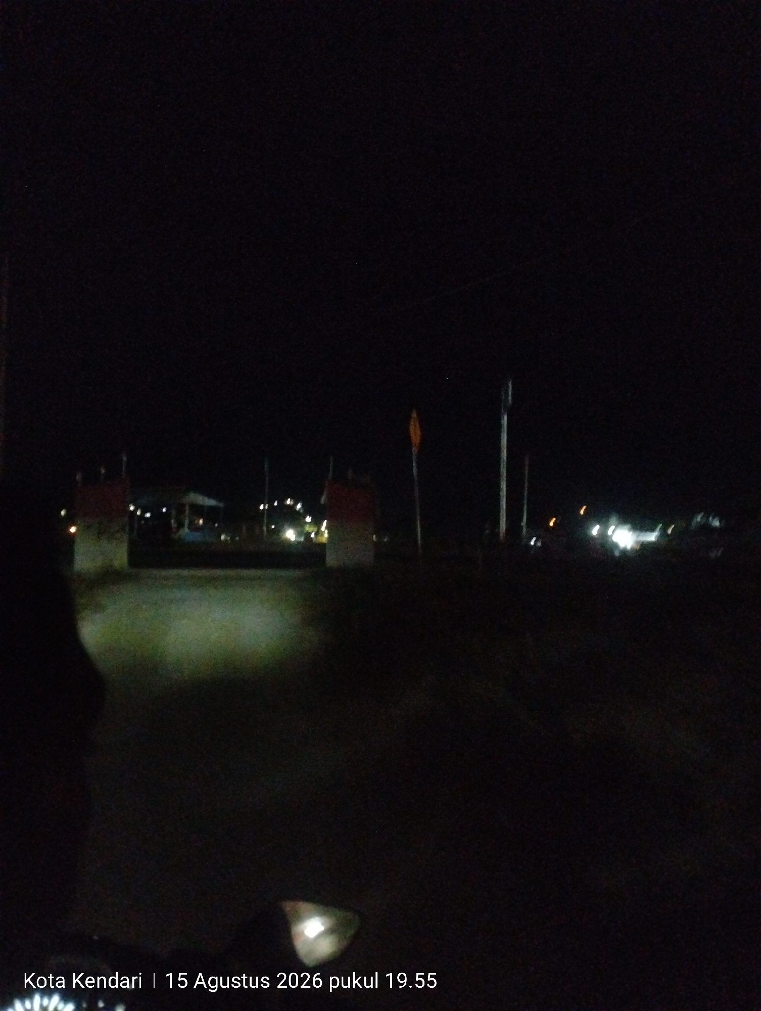 The image is a dark, nighttime photograph taken in Kendari, Indonesia. It appears to be taken from a vehicle, possibly a motorcycle, as evidenced by the headlights illuminating the foreground and the lower portion of the image showing a reflective surface that could be a headlight or a dashboard.
In the mid-ground, there are two distinct structures that resemble archways or gates, painted in red and white. Behind these structures, a building with a roof is visible, along with scattered lights, suggesting a populated area or a facility. Further into the background, more lights are visible, possibly from streetlights or other vehicles. The sky is dark and featureless.
A timestamp at the bottom of the image indicates "Kota Kendari | 15 Agustus 2026 pukul 19.55", confirming the location as Kendari and the date and time as August 15, 2026, at 7:55 PM. The overall scene suggests an outdoor environment at night, possibly on a road leading into or out of a town or a specific location within Kendari.