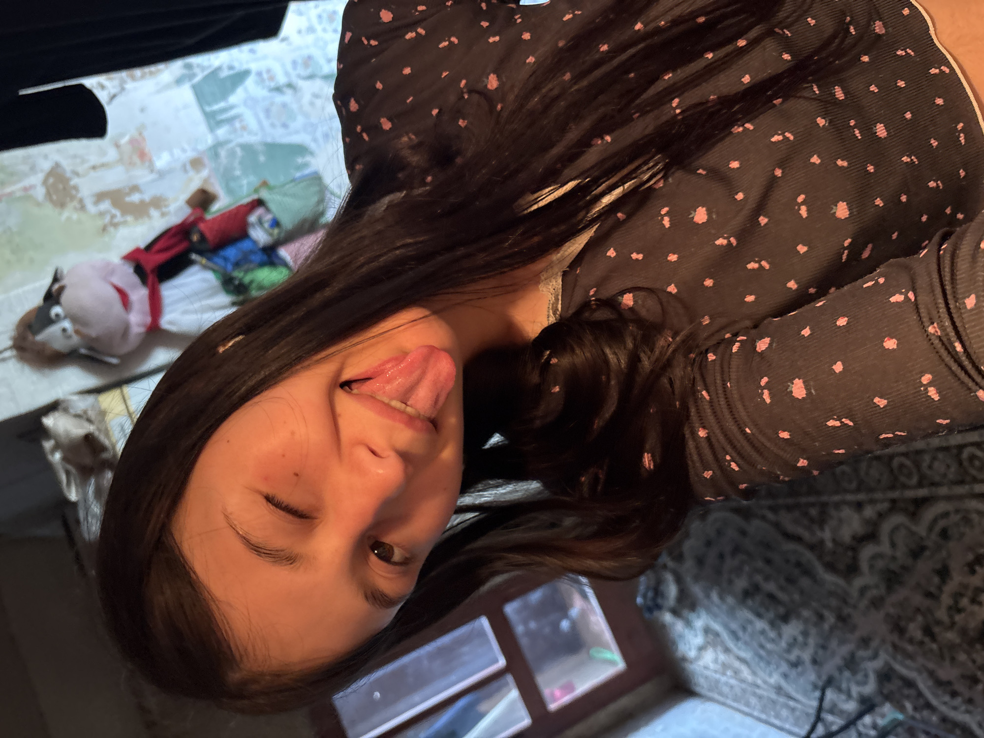 A young person, likely female, is featured in a close-up indoor portrait, playfully winking with one eye and sticking out her tongue. Her dark, long hair cascades over a dark top patterned with small, light pink, abstract dots. The image is oriented on its side, with her head tilted.
In the background, to her left, a light-colored surface with a geometric or tiled pattern is visible. On this surface rests a stuffed animal or puppet with a light body, dark snout, googly eyes, and a red accessory. Other indistinct colorful fabrics are also present. To her right and below, a dark patterned rug or carpet can be seen. On a darker surface near her face, several clear plastic containers are visible, with one holding small pink and green objects. The scene is illuminated by indoor artificial lighting, creating a lighthearted atmosphere. No text is visible in the image. The location context is Фоки, Russia.